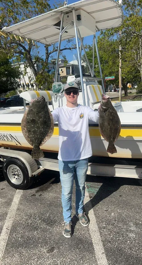 A person holding two large flounder fish in front of a boat, with trees in the background.