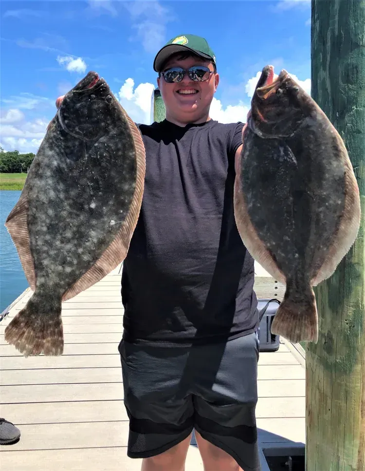 Person on a dock holds two large, speckled flounder; sunny day.