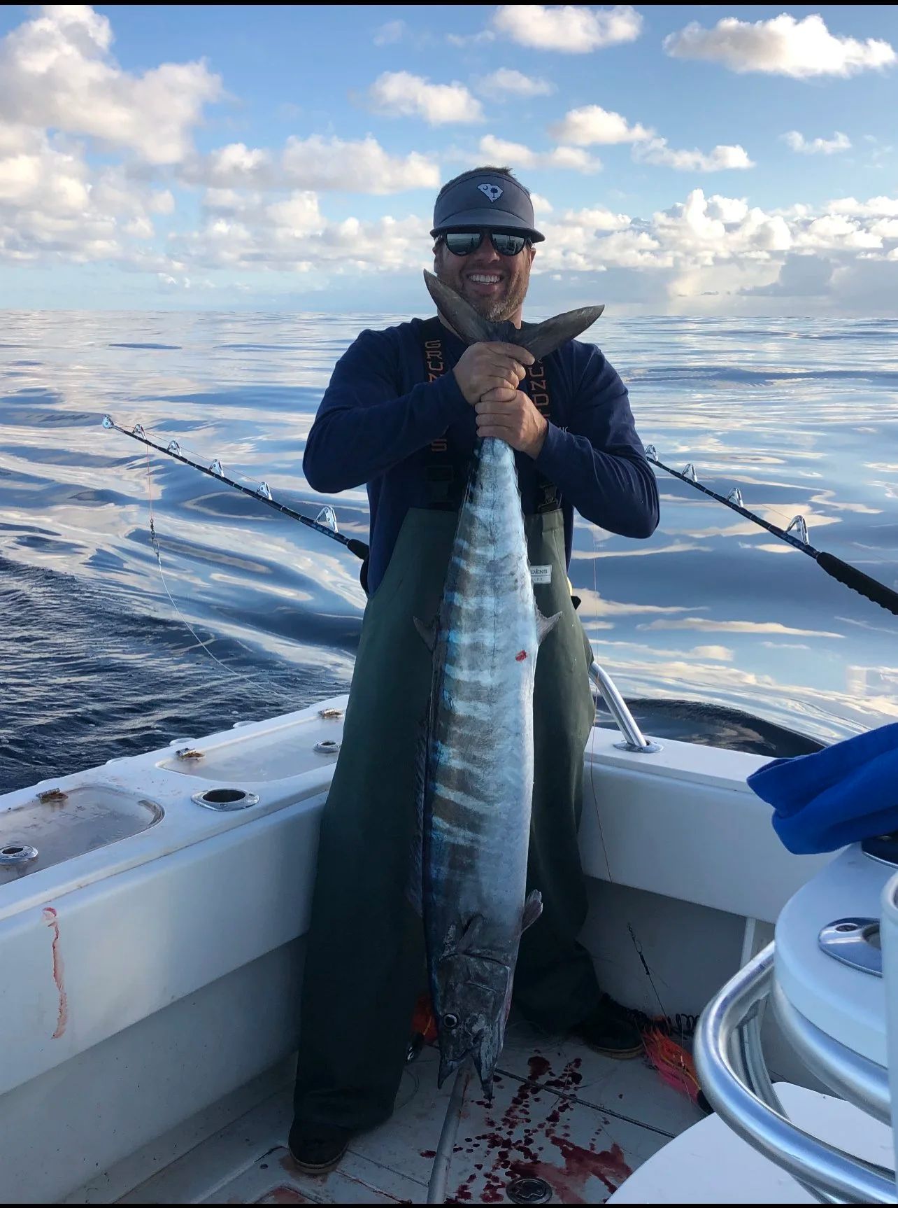 Man on a boat smiles while holding a large fish, with ocean and cloudy sky in background.
