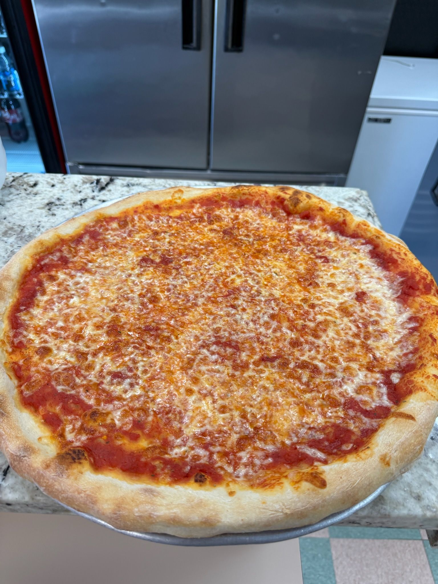 Cheese pizza on a metal tray in a kitchen, with a browned crust and tomato sauce.