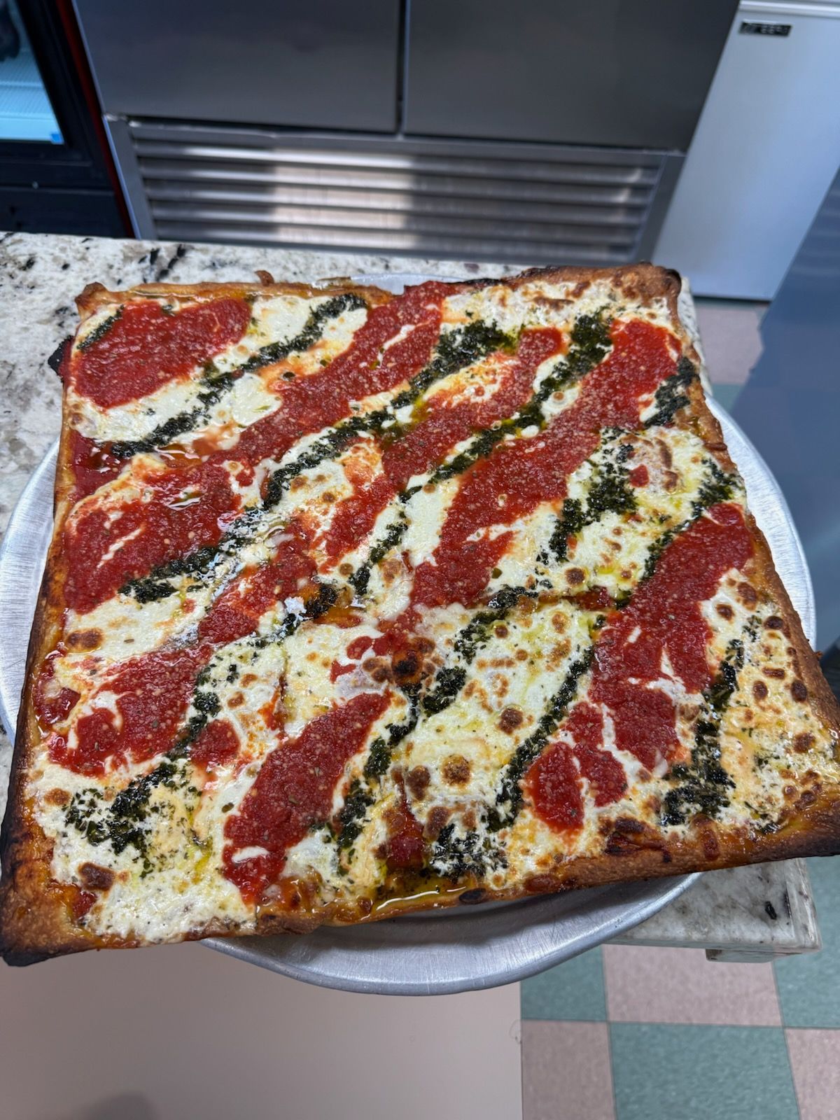 Square pizza on a tray with red sauce and white cheese, cut into slices on a kitchen counter.