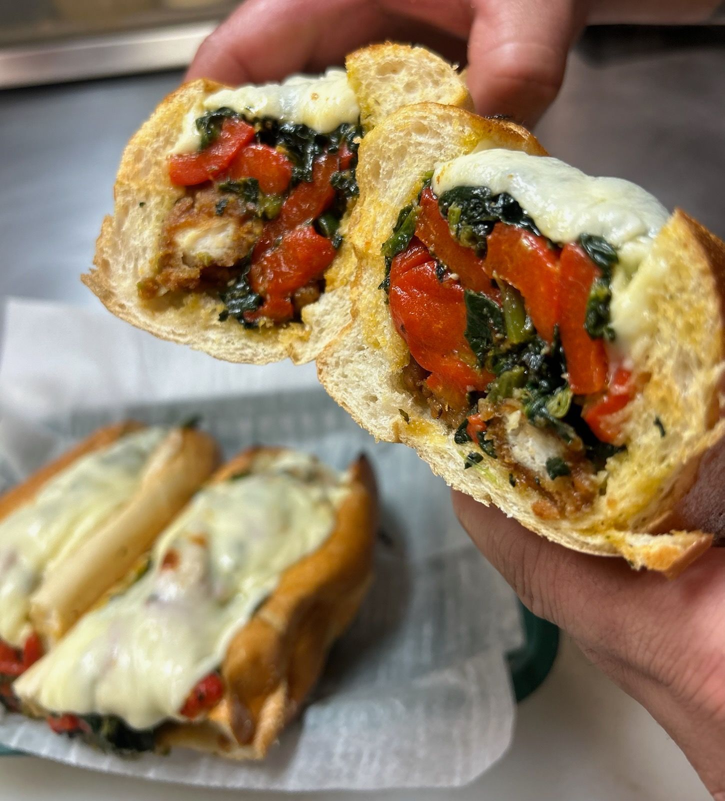 Hand holding a stuffed sandwich with melted cheese, roasted peppers, and greens; another sandwich on paper below