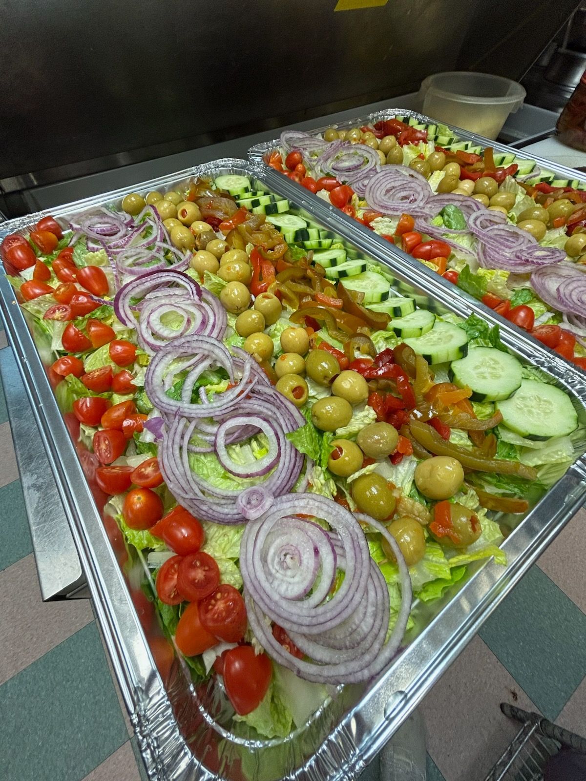 Tray of mixed salad with sliced onions, tomatoes, cucumbers, and olives in a buffet line