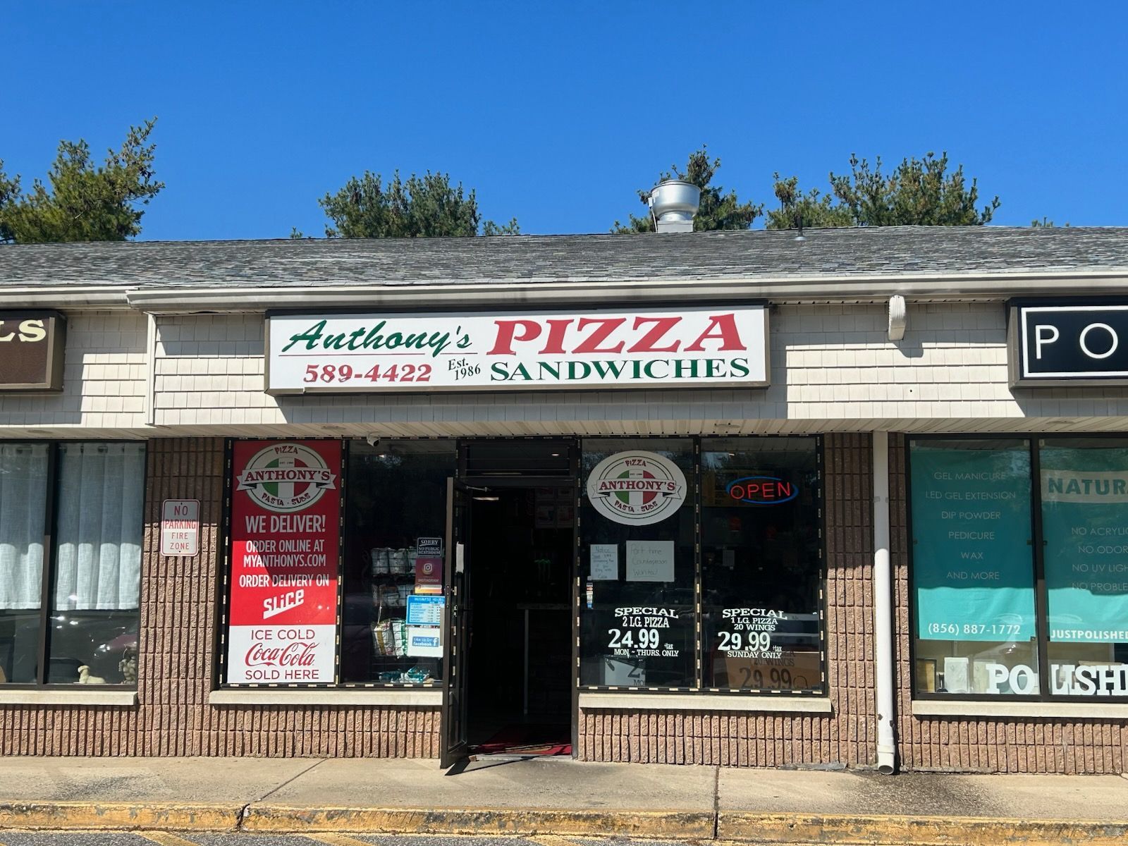 Strip mall storefront for Anthony's Pizza and Sandwiches with red-and-green sign and open entrance.