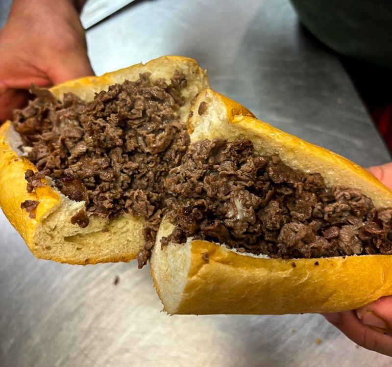 A hand holds a sandwich stuffed with chopped beef on a metal counter.