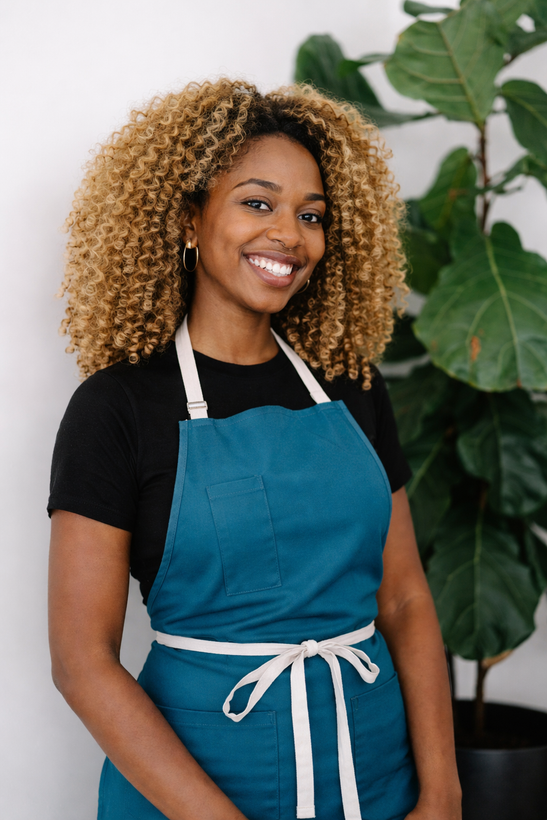 A smiling person wearing a dark teal apron over a black shirt, standing in front of a tall green plant against a white wall.