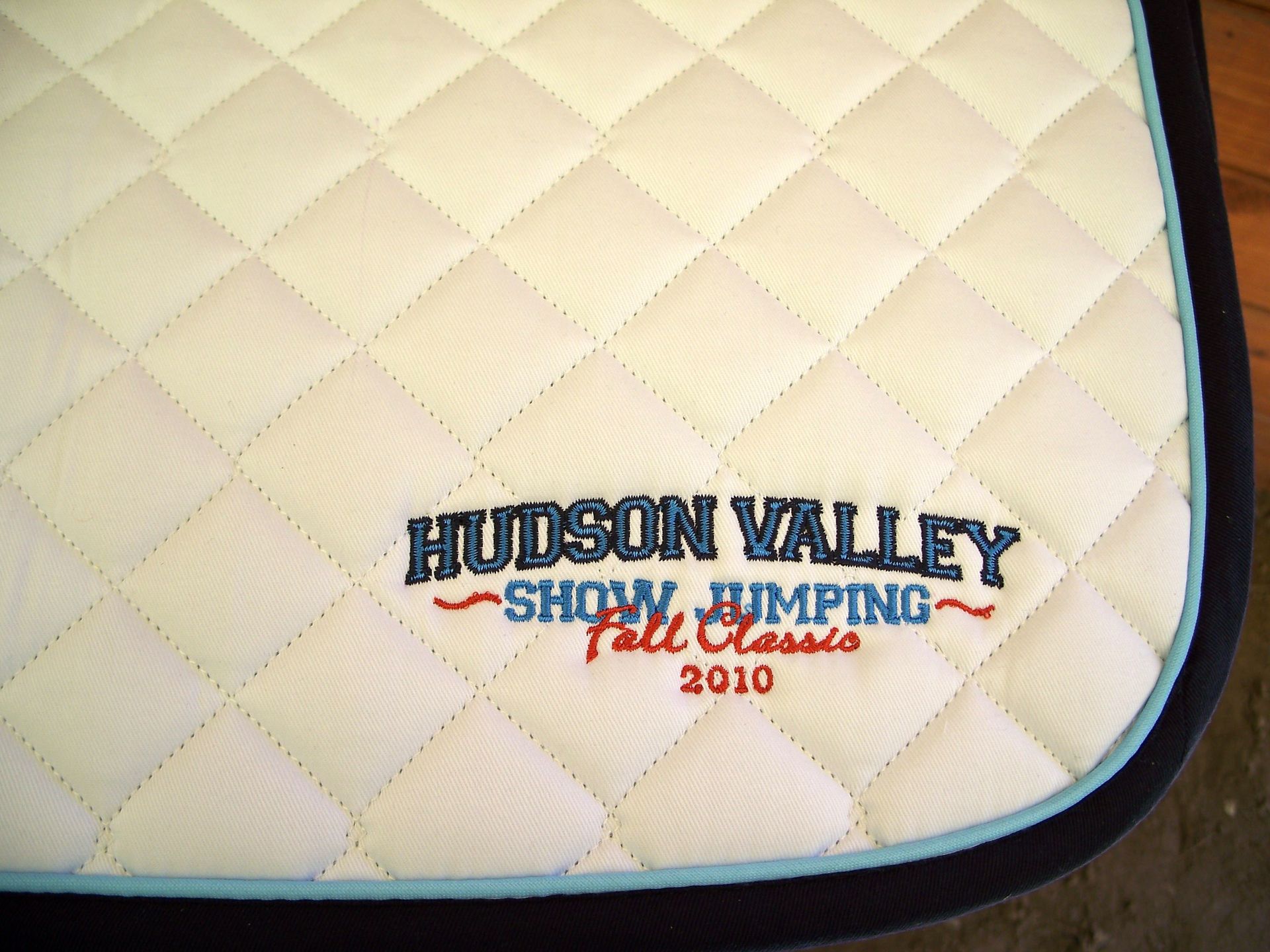 A quilted white horse saddle pad with a light blue and dark blue trim, embroidered with 