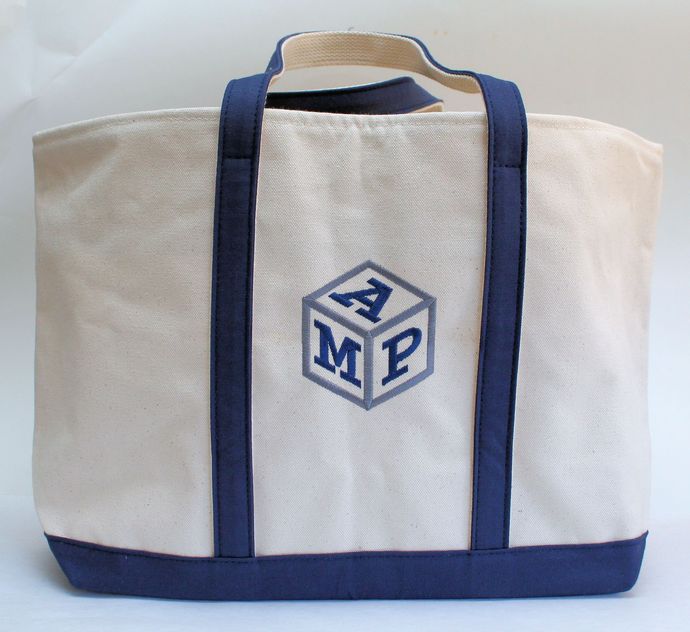 A cream-colored canvas tote bag with navy blue straps, base, and an embroidered cube monogram featuring the letters AMP.