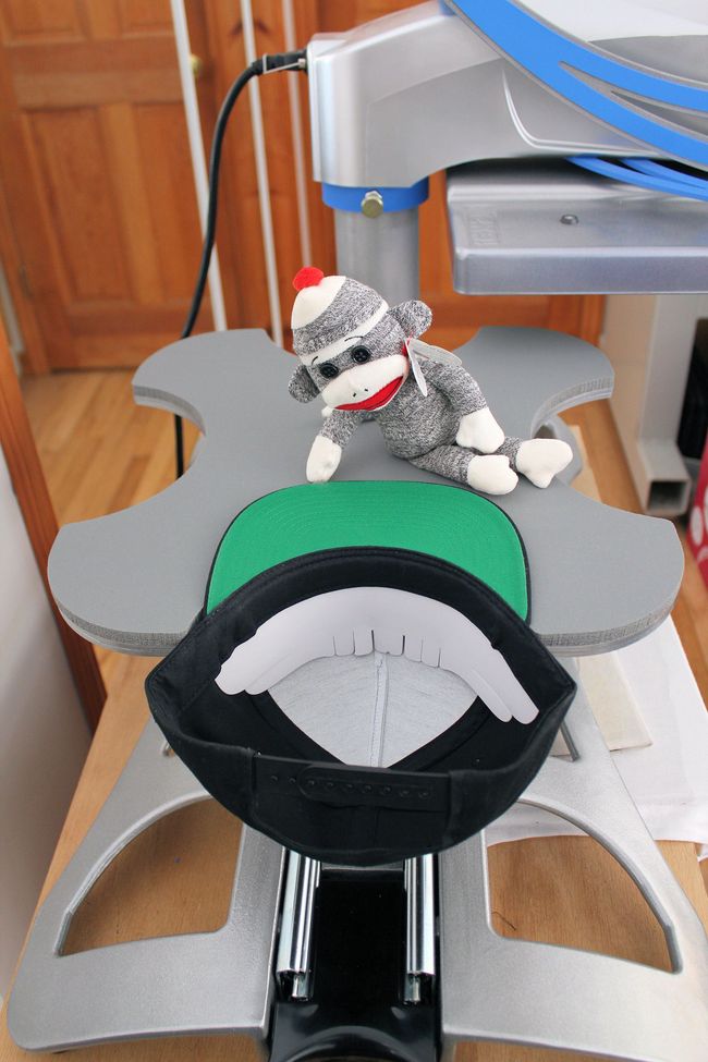 A sock monkey toy sits on a grey heat press machine next to a black baseball cap with a green brim.