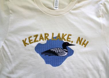A cream-colored t-shirt with an embroidered blue lake shape, a black and white loon, and the text 