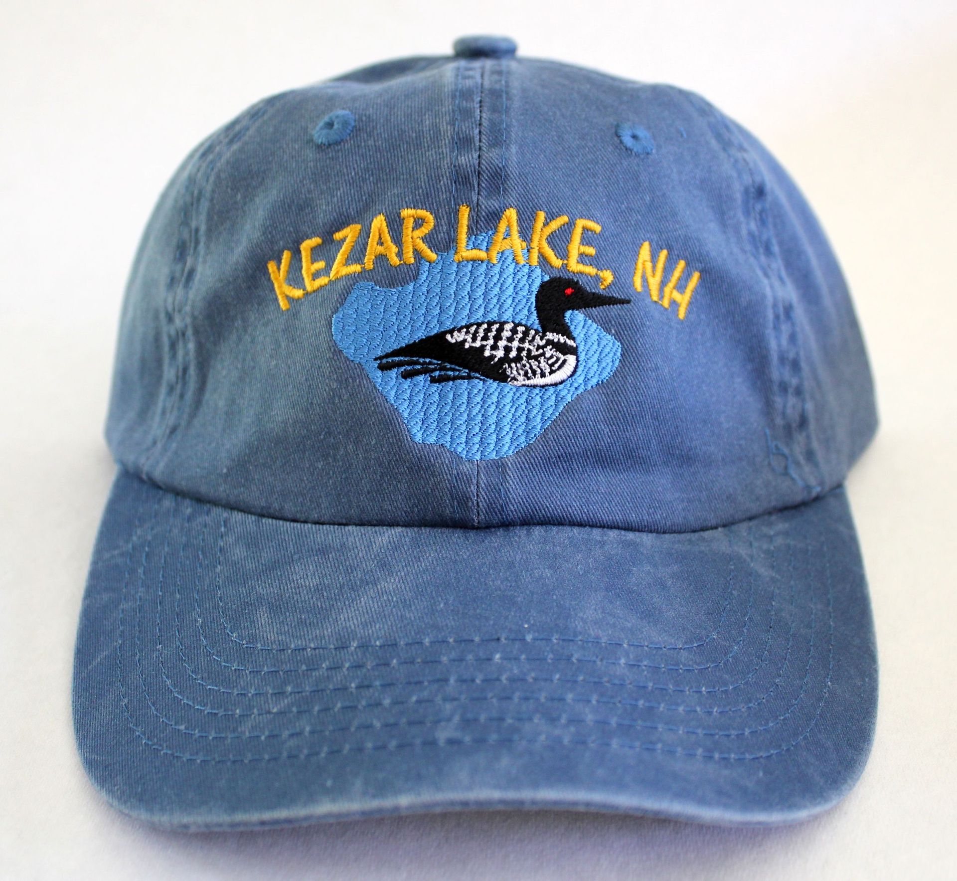 A blue baseball cap with an embroidered design of a loon on a blue lake shape and yellow text reading A blue baseball cap with an embroidered design of a loon on a blue lake shape and yellow text reading