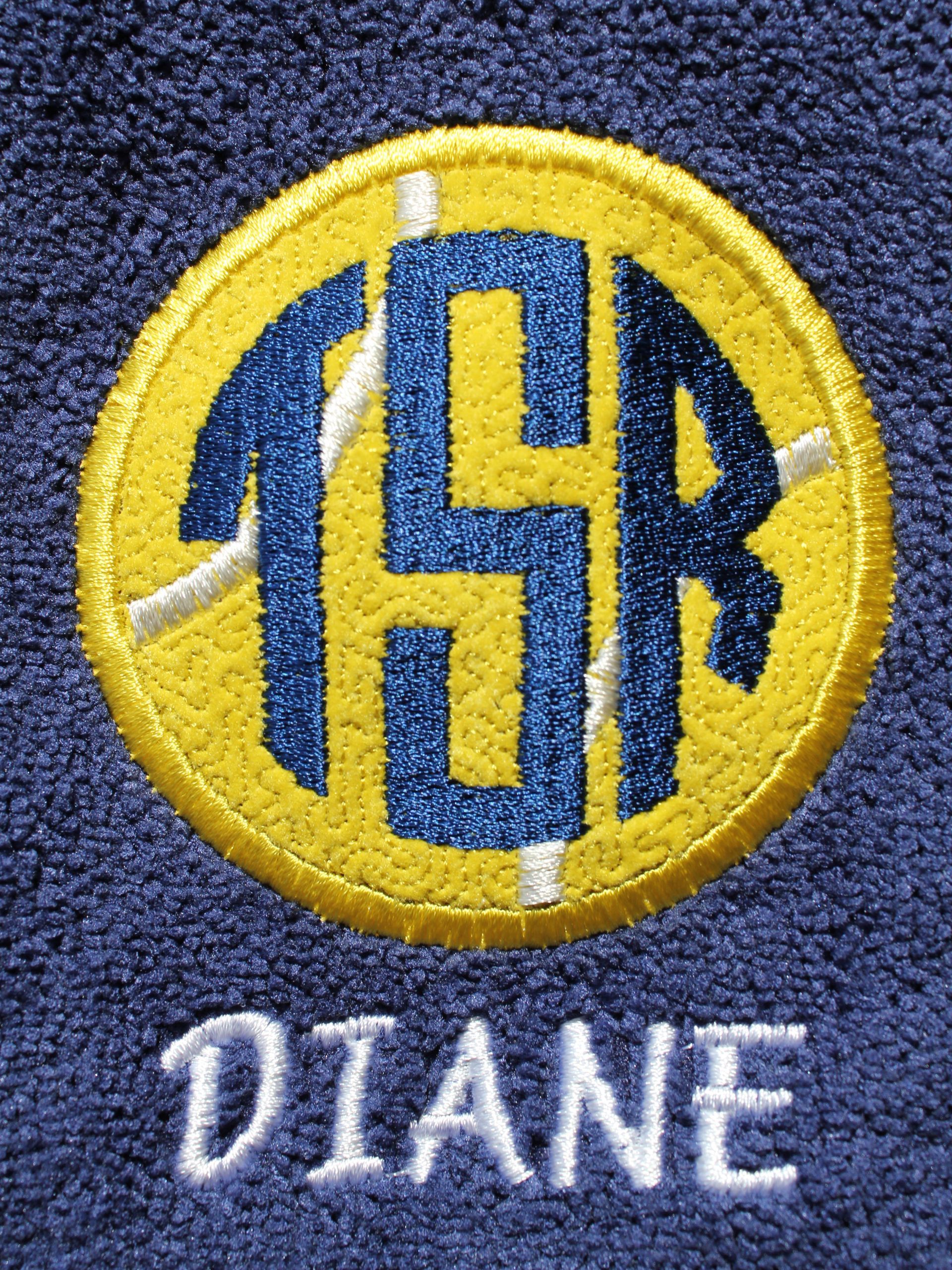 A navy blue fabric embroidered with a circular yellow patch featuring the dark blue monogram 