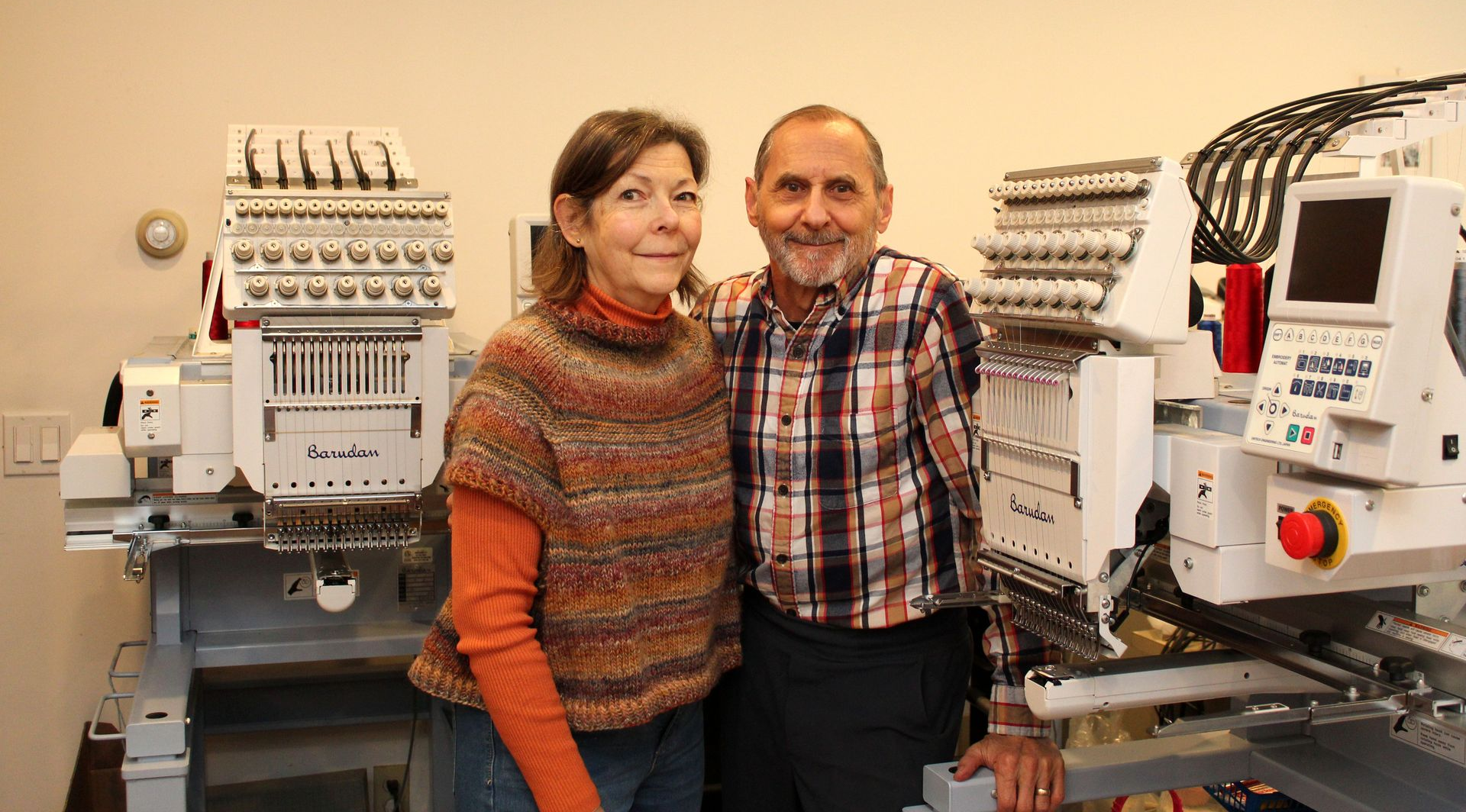 A person in a knit sweater and a person in a plaid shirt stand smiling between two industrial embroidery machines.