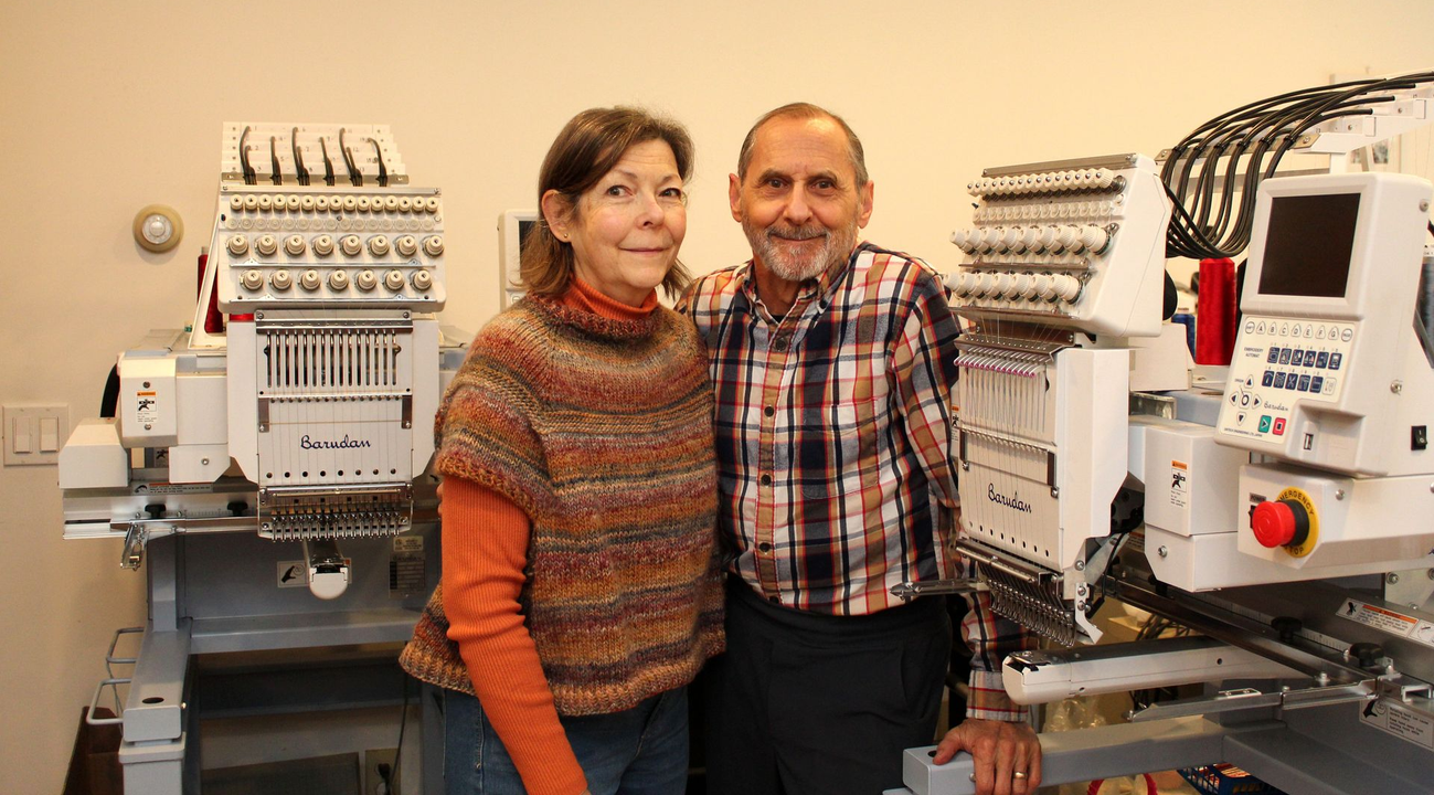 A man and a woman stand smiling between two industrial embroidery machines in a room with light-colored walls.