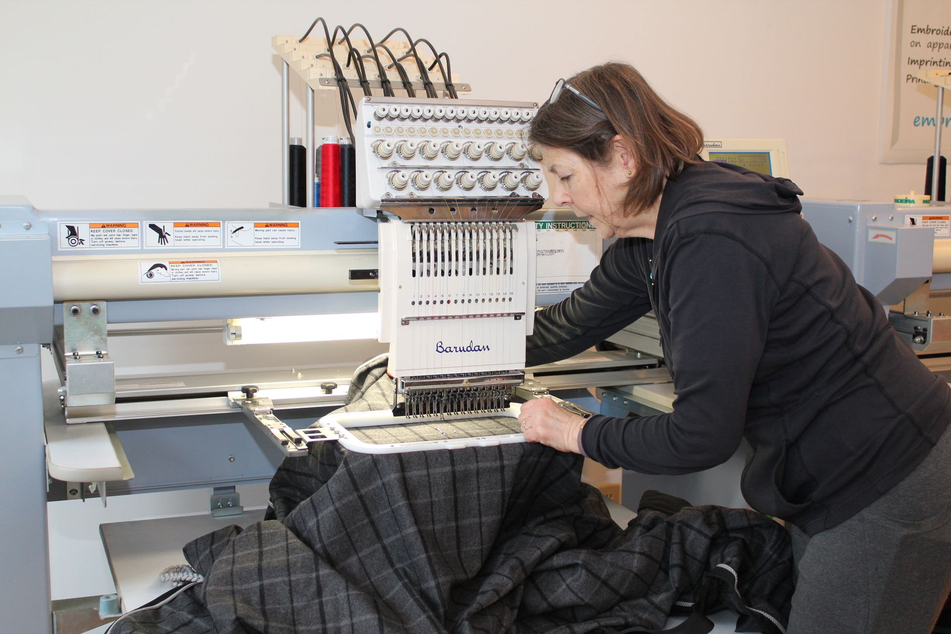 A person using an industrial embroidery machine on a piece of dark plaid fabric in a workshop setting.