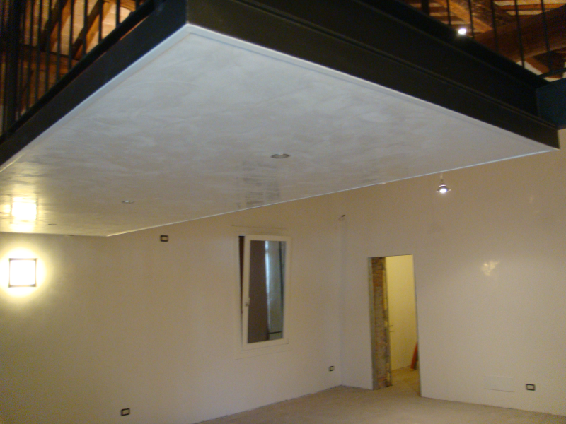 Un soppalco con struttura in metallo nero si protende su una stanza con pareti bianche, una piccola finestra rettangolare e un'apertura per la porta.