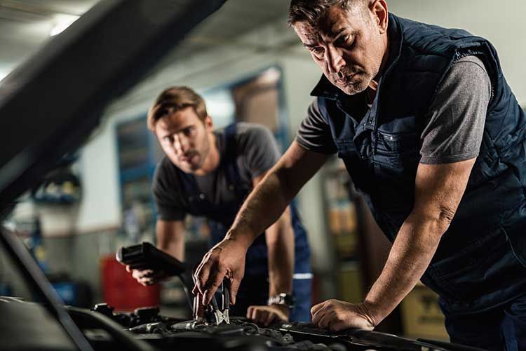 Two auto mechanics inspecting a car engine in a garage. One holds tools, the other a diagnostic device. | Elite Autocare of Princeton