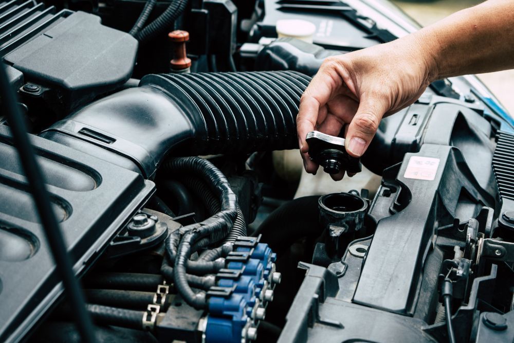 Person's hand removing a cap from a car engine's radiator. Black engine components are visible. | Elite Autocare of Princeton