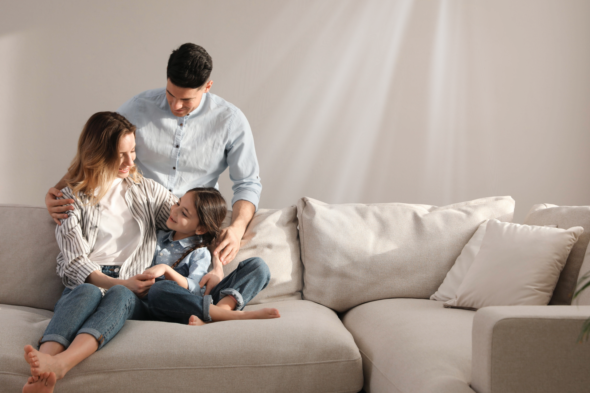Family of three on a light-colored couch, smiling. Sunlight beams on the wall.