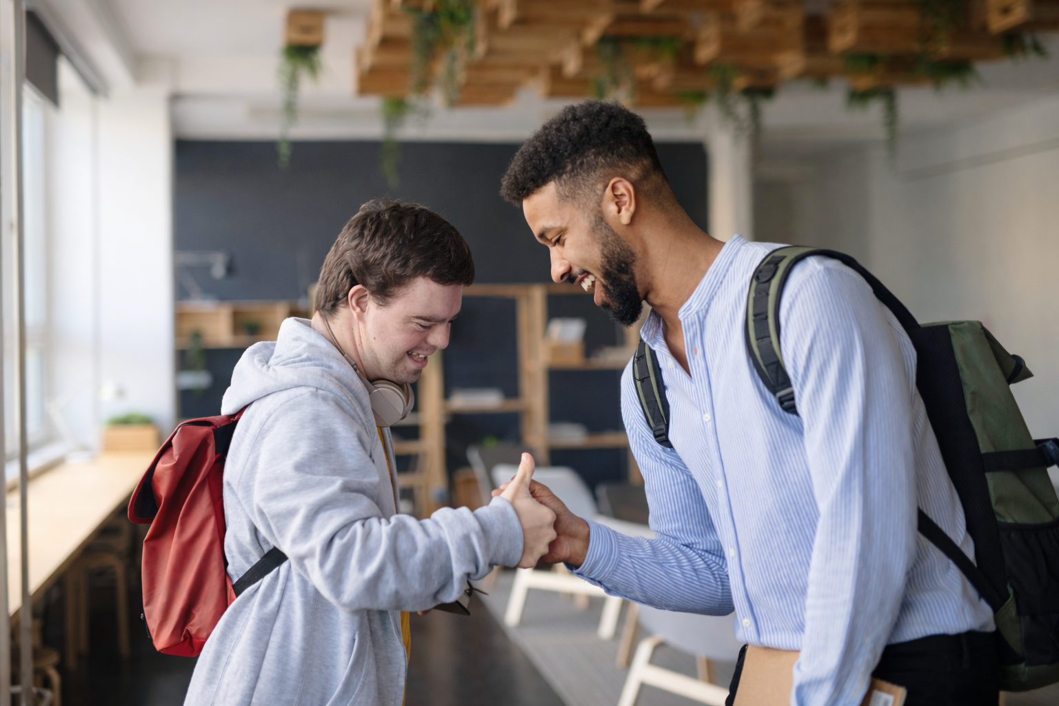 Two people in a hallway, smiling and shaking hands, one with a red backpack, who has Down syndrome and one with a black backpack.