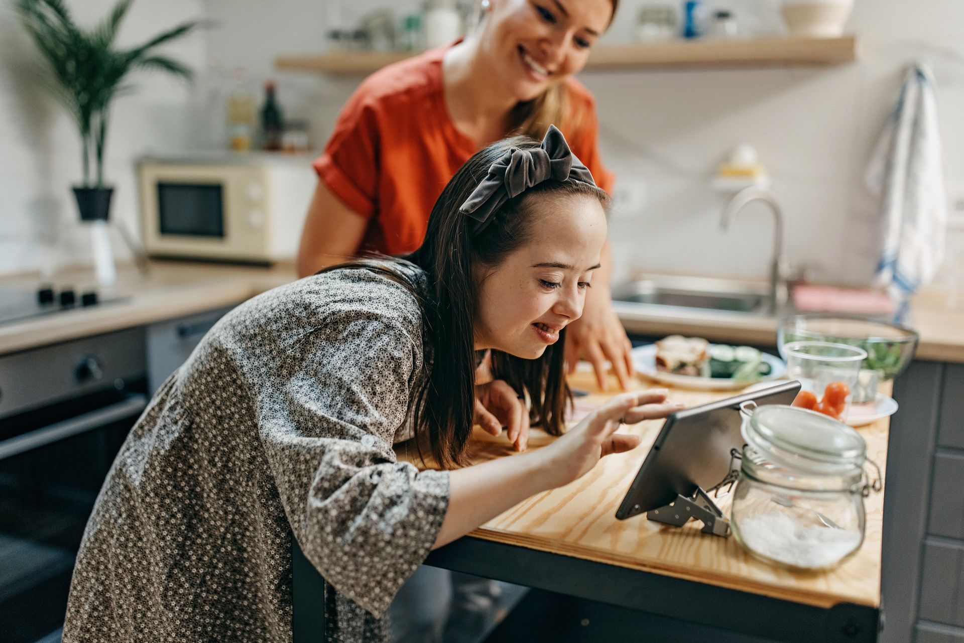 Two people are preparing food at a kitchen table, with jars, bowls, and a laptop nearby.