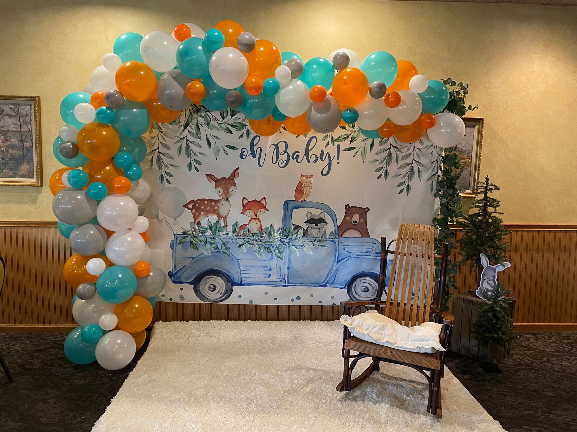 A baby shower backdrop featuring a woodland animal theme, blue truck illustration, balloon arch, and a wooden rocker.