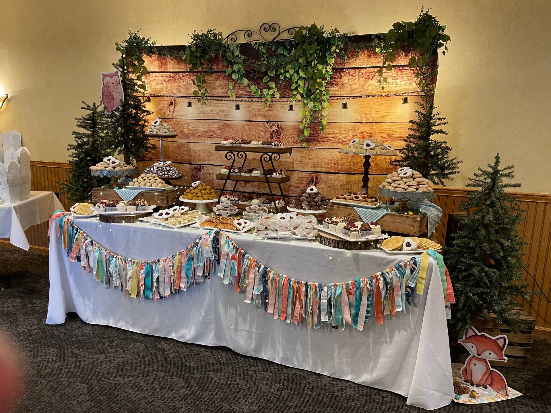 Dessert table with treats, rustic backdrop, garland, and small trees. Fox decoration on the floor.