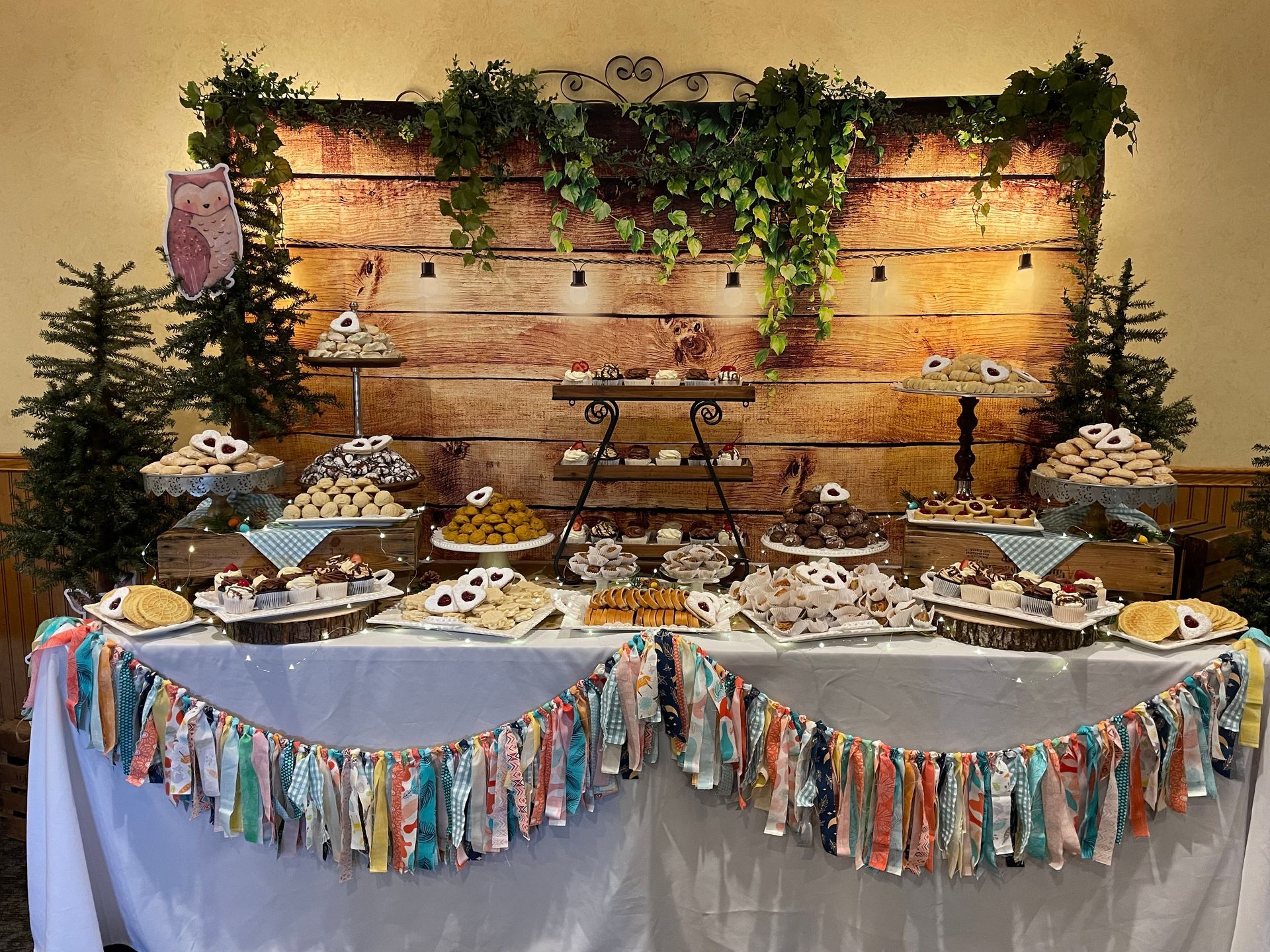 A dessert table featuring tiered trays of treats, festive greenery, string lights, and a colorful fabric banner backdrop.