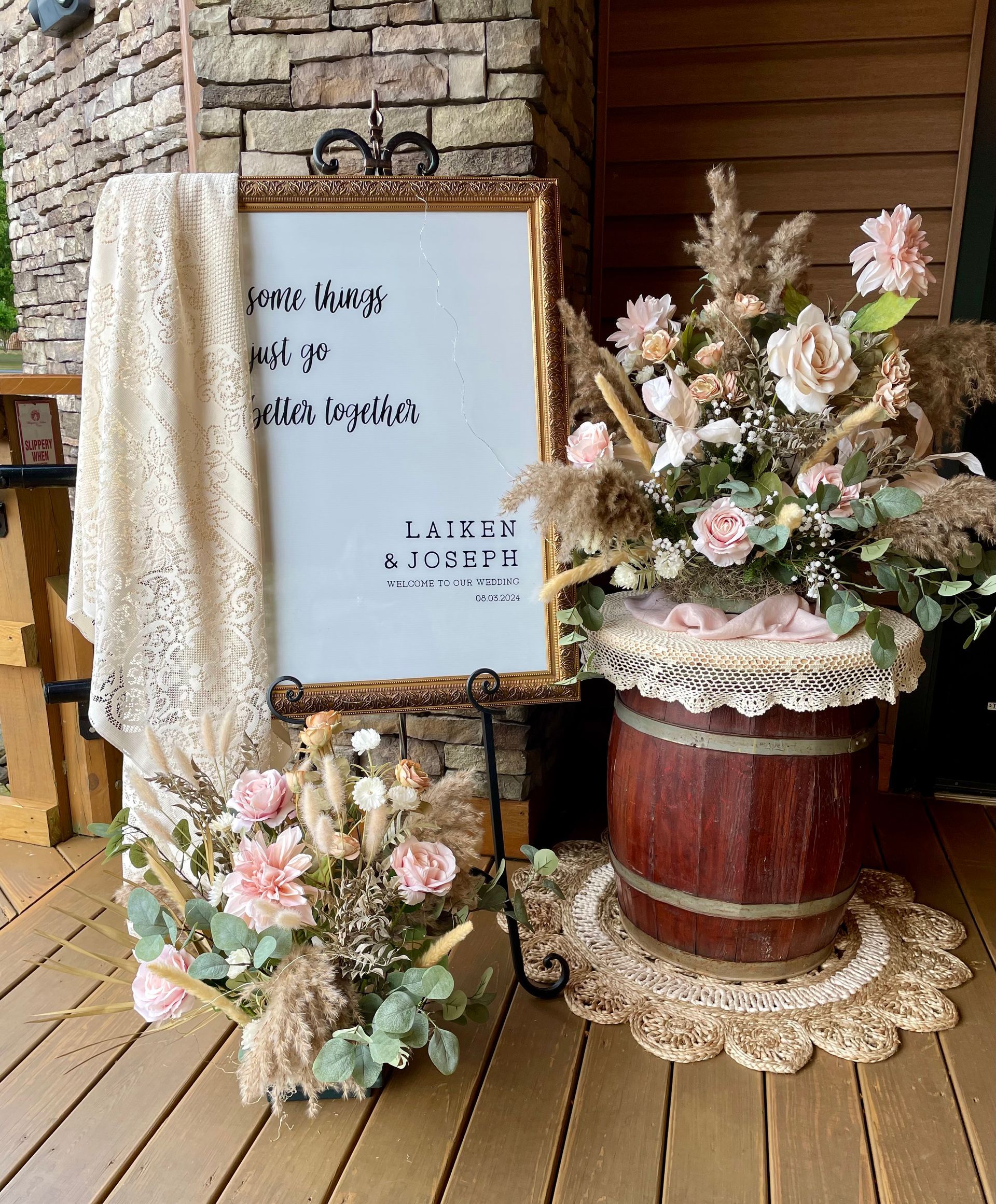 A sign on an easel with a lace cloth, floral arrangements, and a wooden barrel, decorated for a wedding event.