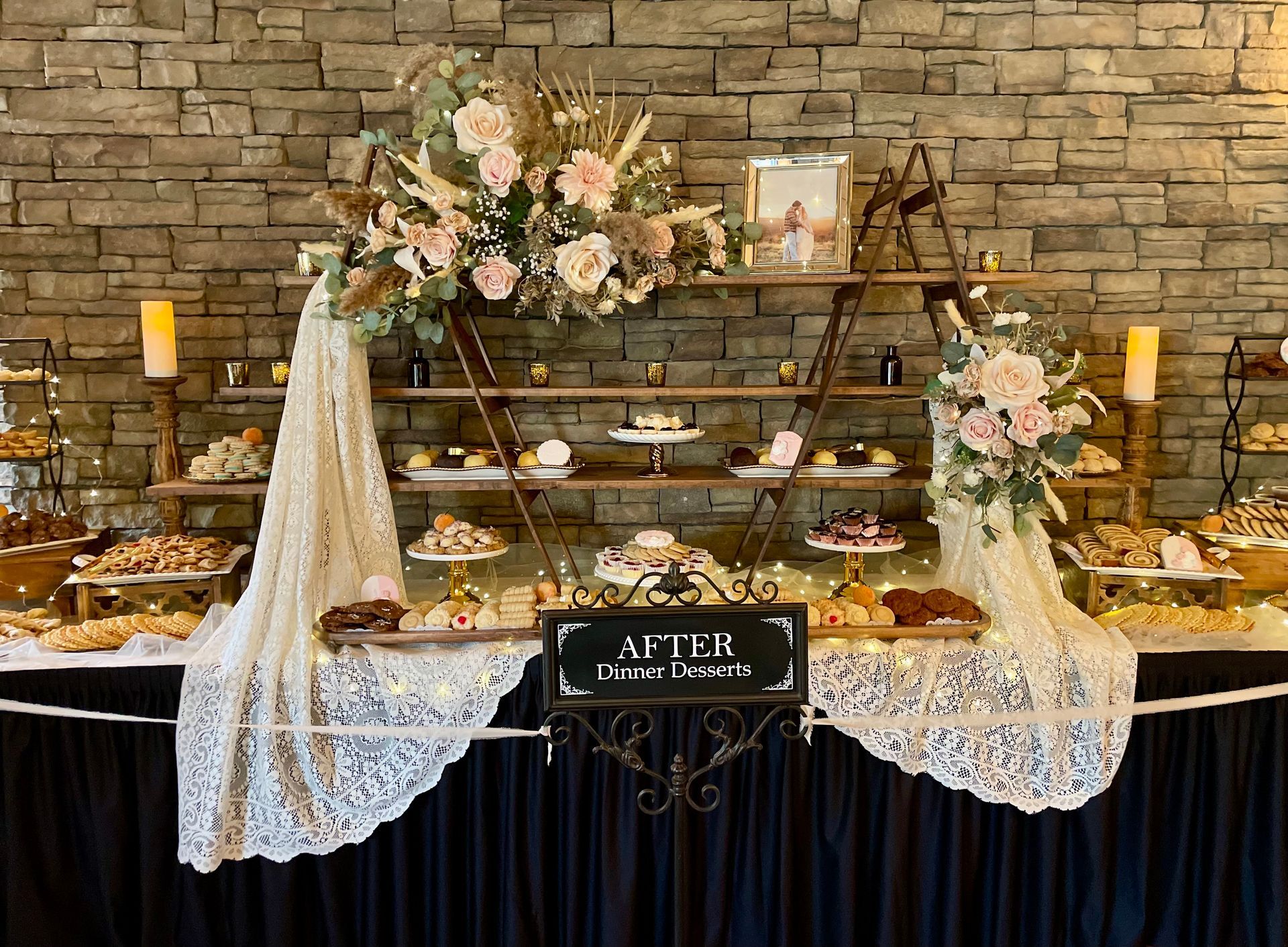 A dessert table with a tiered wooden stand, lace draping, floral arrangements, and a sign labeled 
