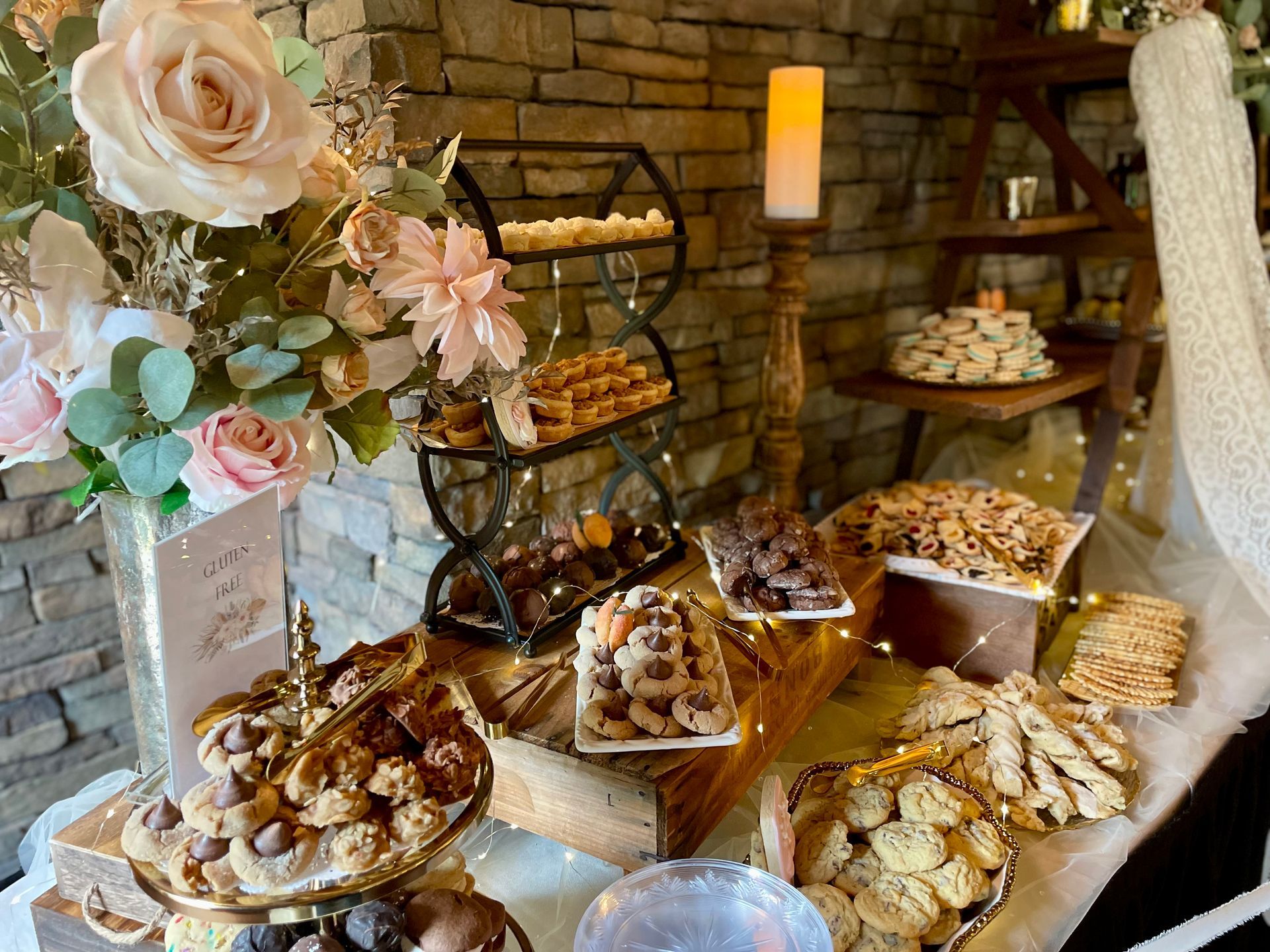 A dessert table with tiered trays of cookies and pastries, decorated with flowers and a candle in a rustic, stone setting.