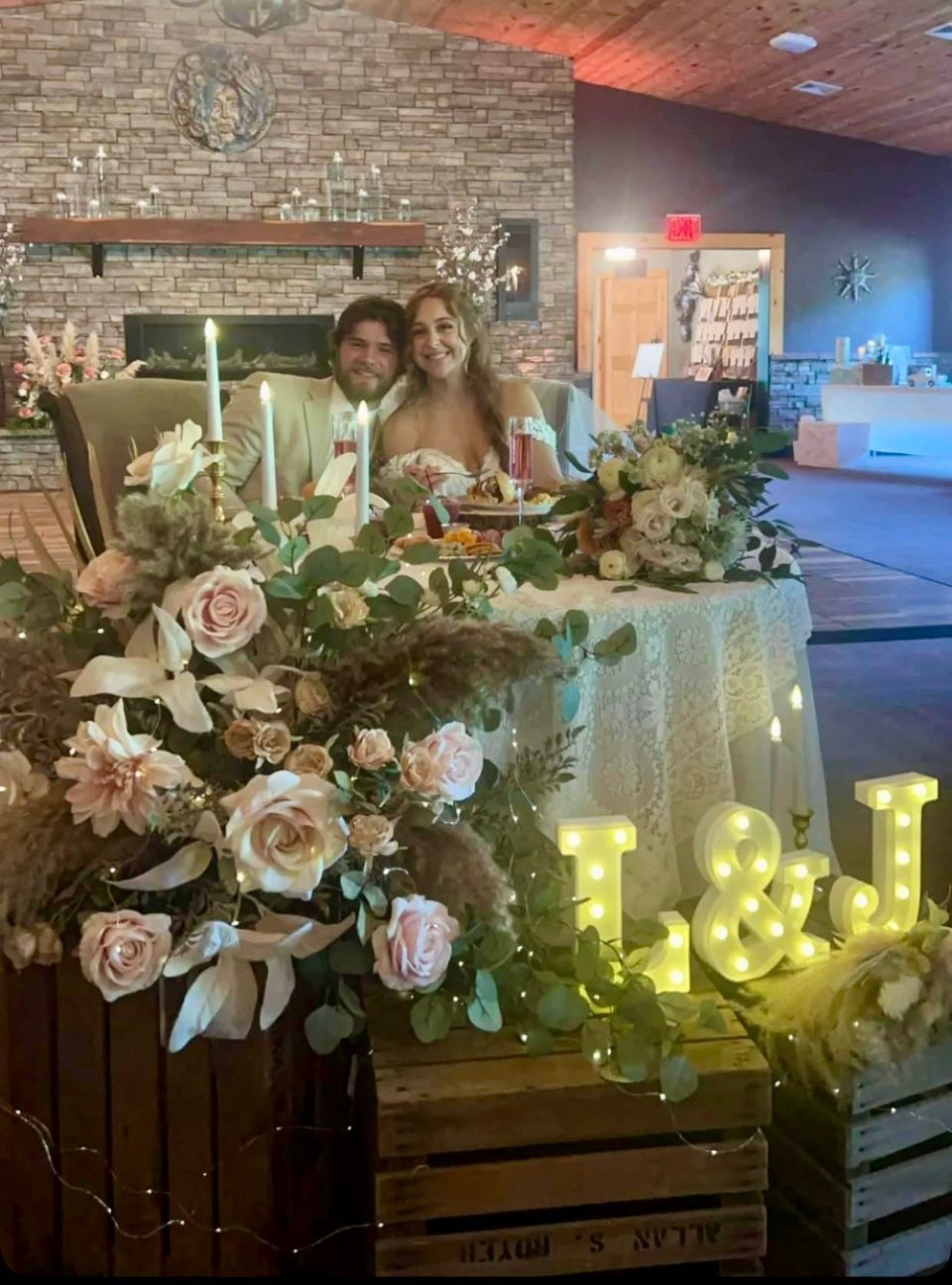 A couple sits at a decorated wedding table with floral arrangements, glowing lights, and a stone fireplace in the back.