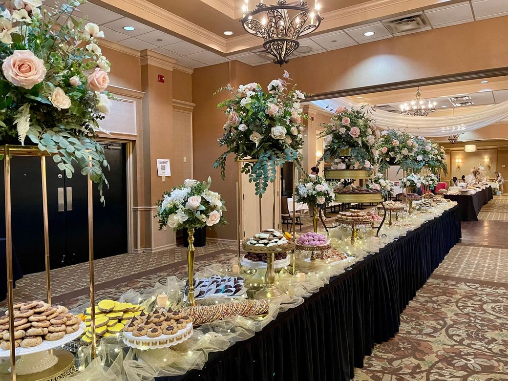 A long banquet table set with floral centerpieces, varied desserts, and treats in a carpeted event hall.