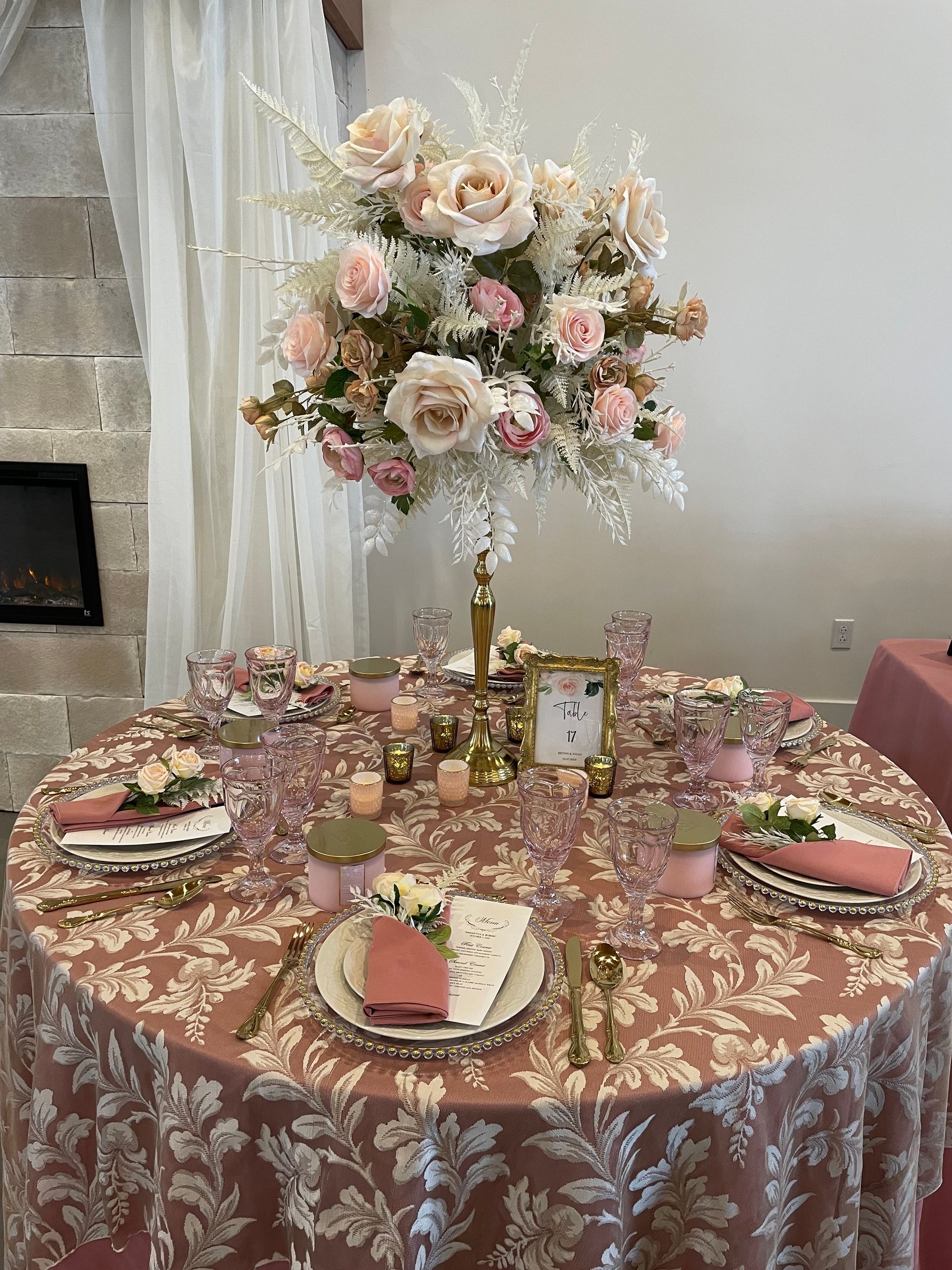 A formal table setting with a pink floral tablecloth, a tall rose centerpiece, candles, and place settings.