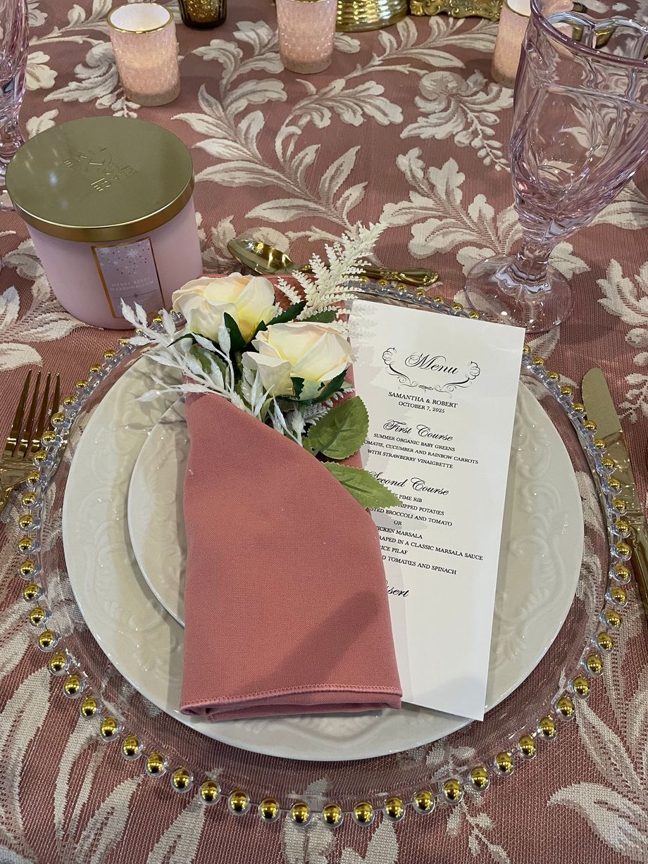 Formal place setting: pink napkin and flowers on white plates, menu, gold accents.