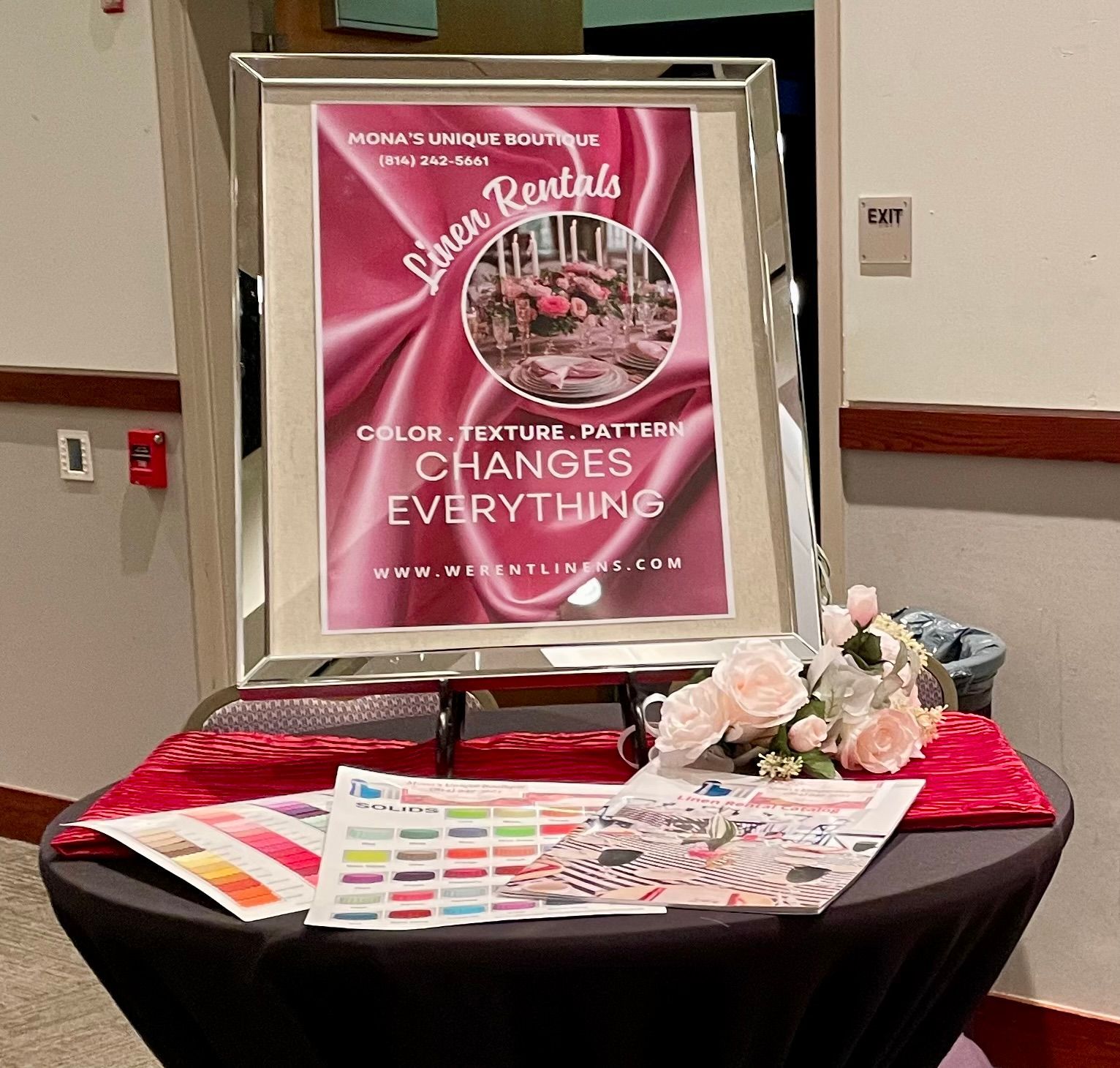 A framed sign for "Moma's Unique Boutique" linen rentals rests on a table decorated with flowers and sample color charts.