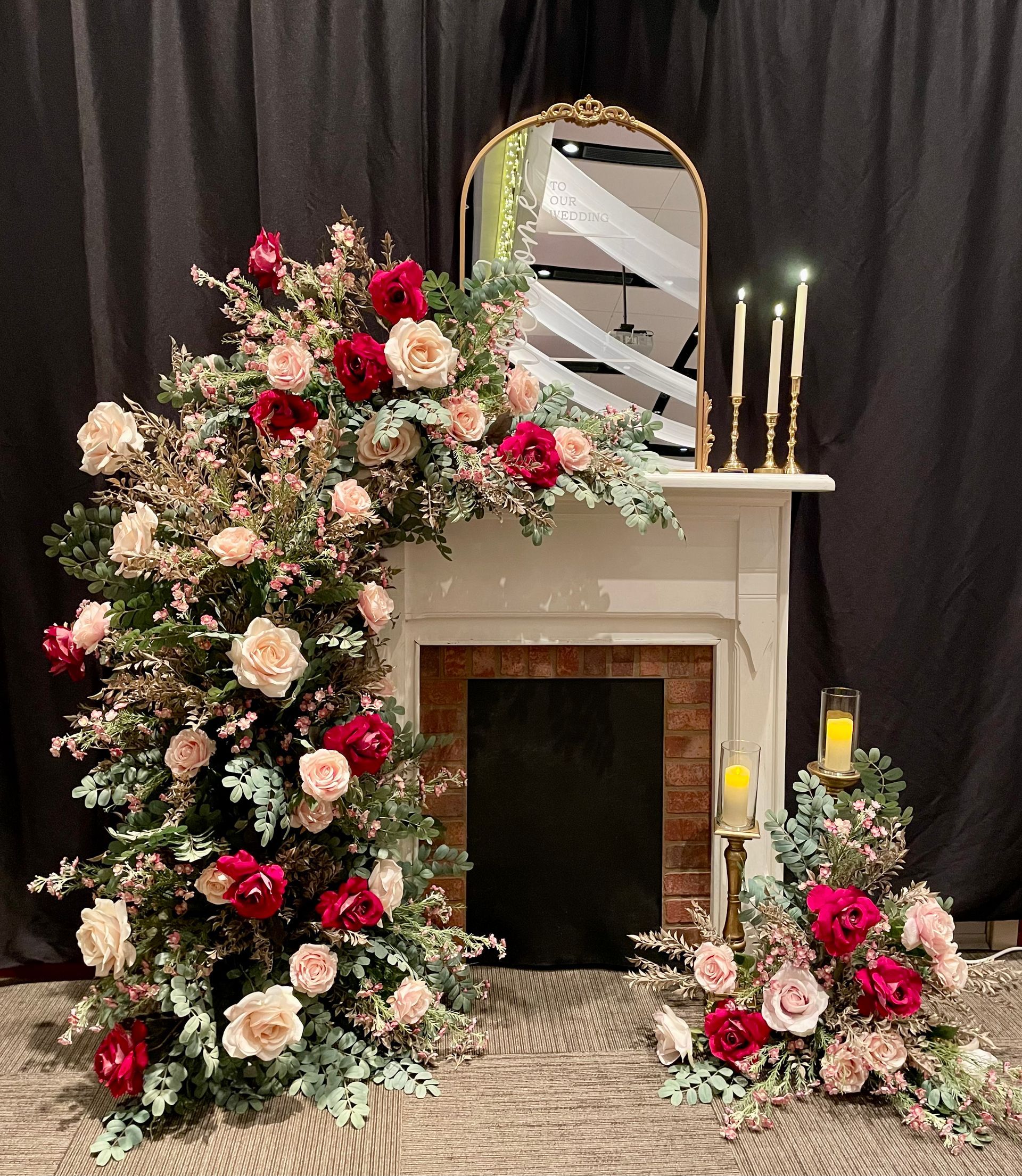 A white faux fireplace mantel decorated with lush pink and red roses, a gold mirror, and glowing candles on a dark stage.