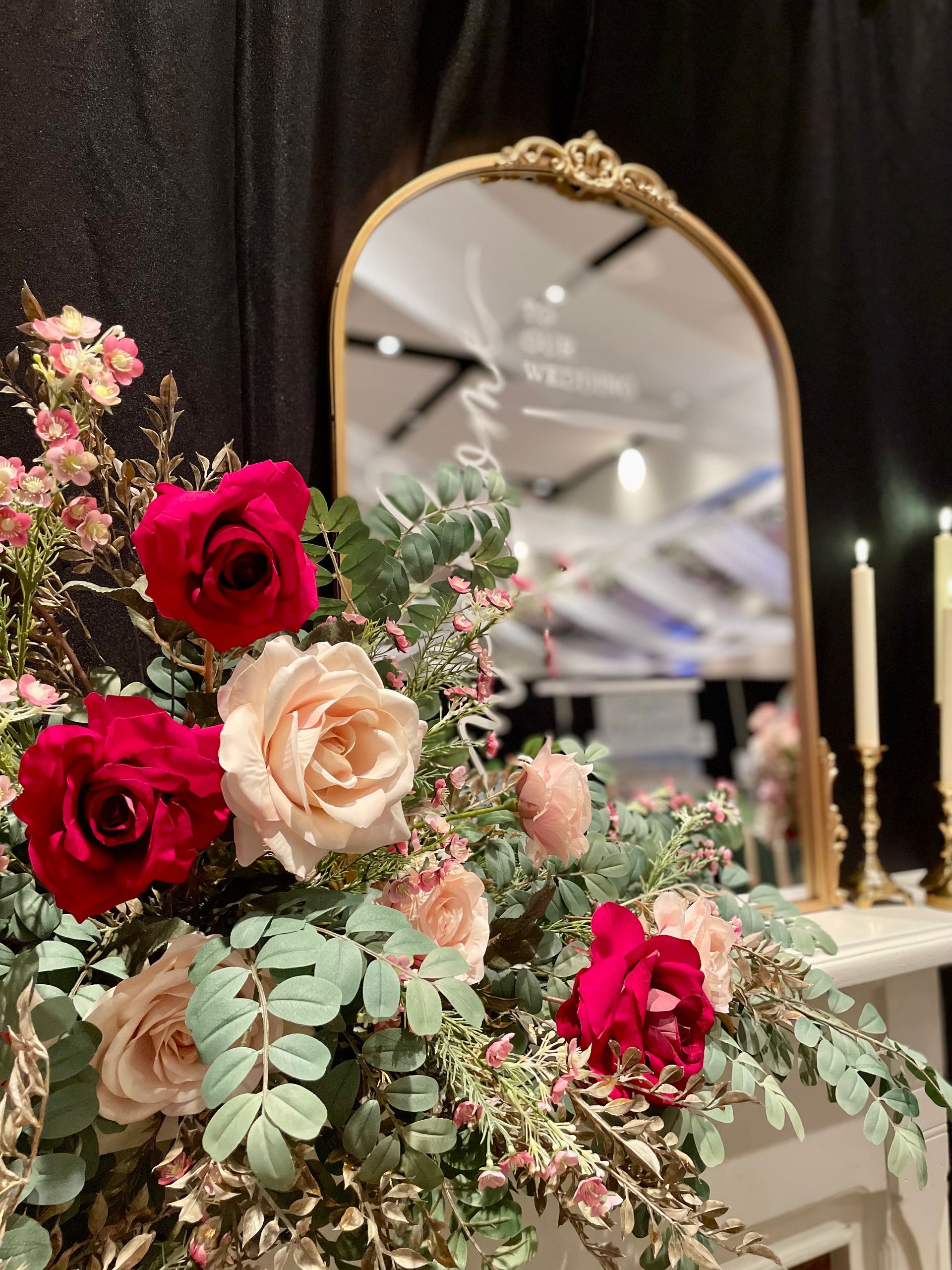 A floral arrangement of red and pale pink roses with greenery stands before an arched gold mirror on a white table.