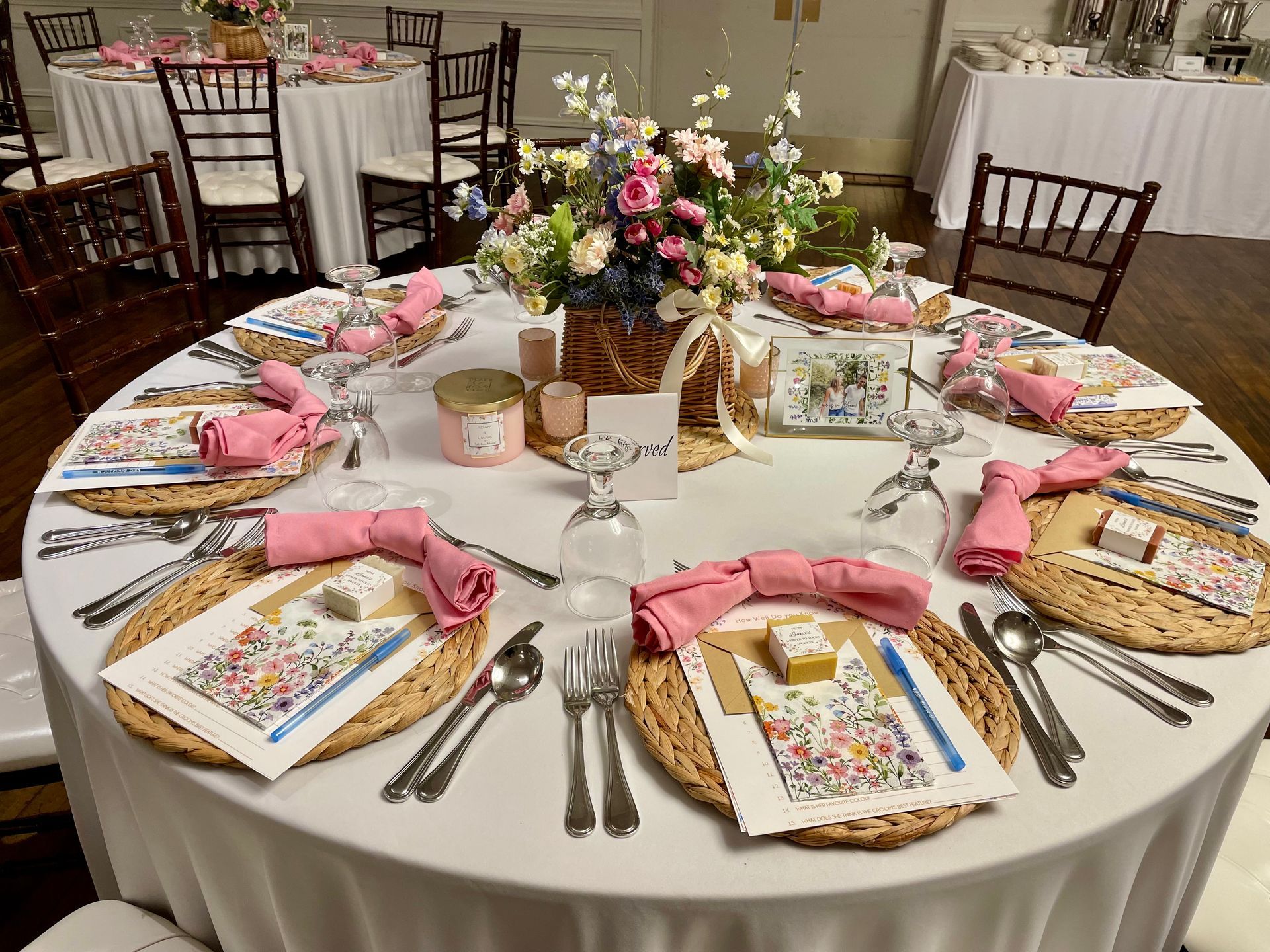 A round event table set with white tablecloth, woven chargers, pink napkins, floral place cards, and a central centerpiece.