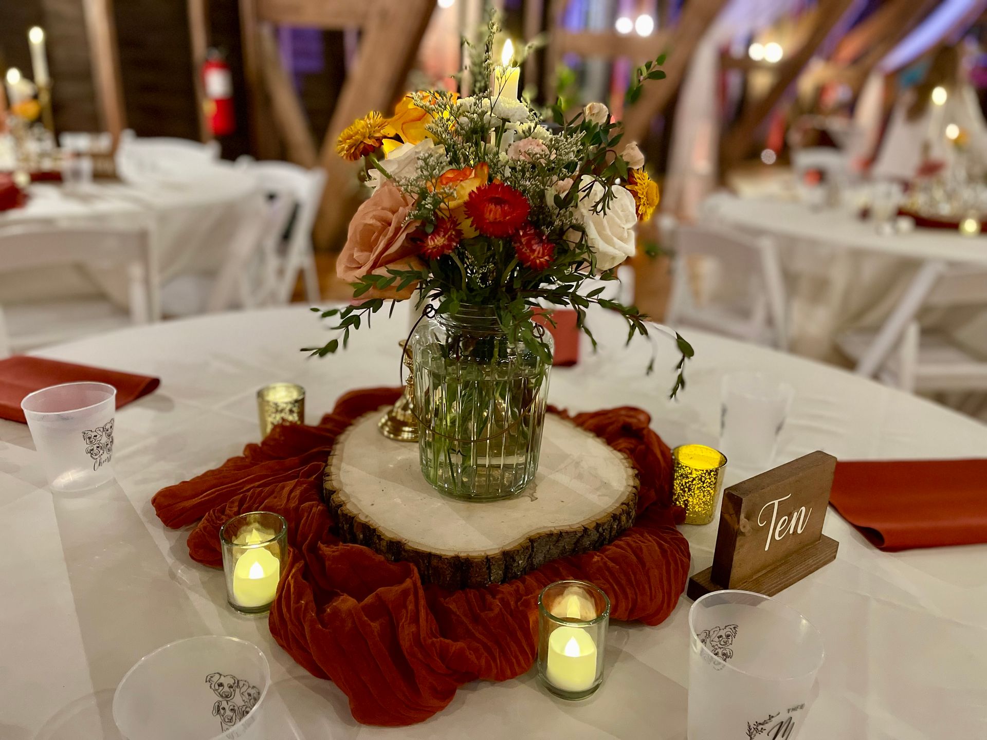A rustic wedding centerpiece with a floral mason jar on a wood slice, surrounded by red fabric and small lit candles.