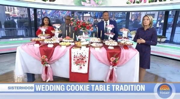 Four people stand behind a table filled with assorted cookies, decorated with pink and red cloths, on a news set.