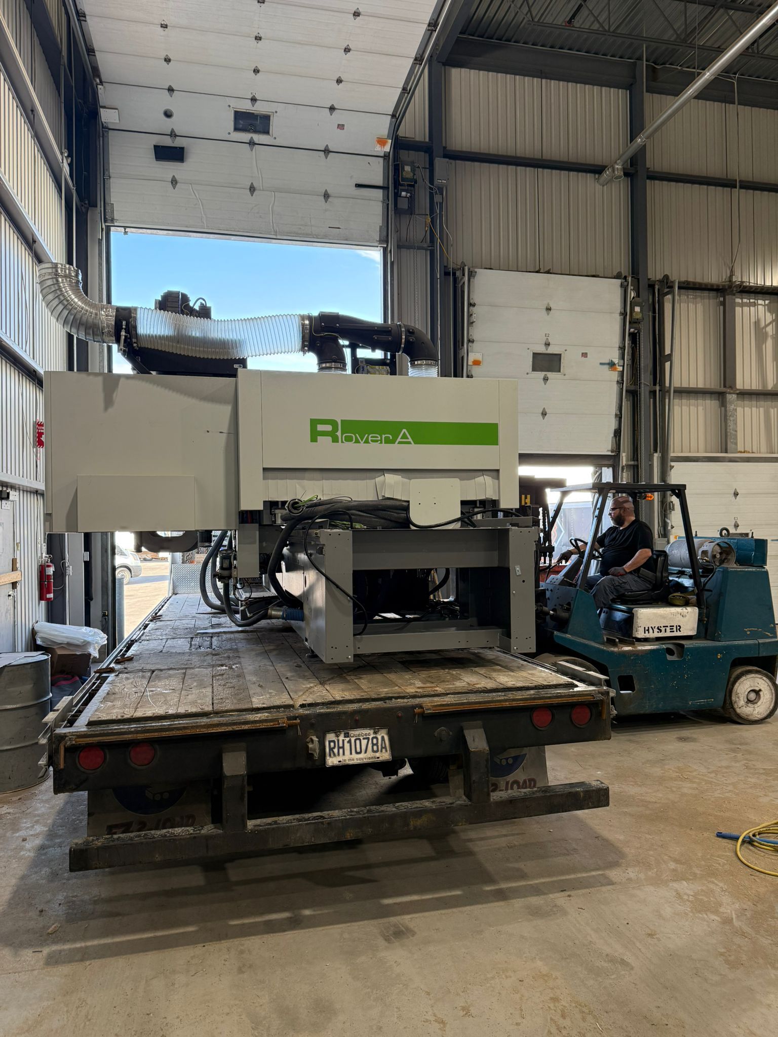 A large, gray industrial machine is on a flatbed truck, being moved with a forklift, in a warehouse.
