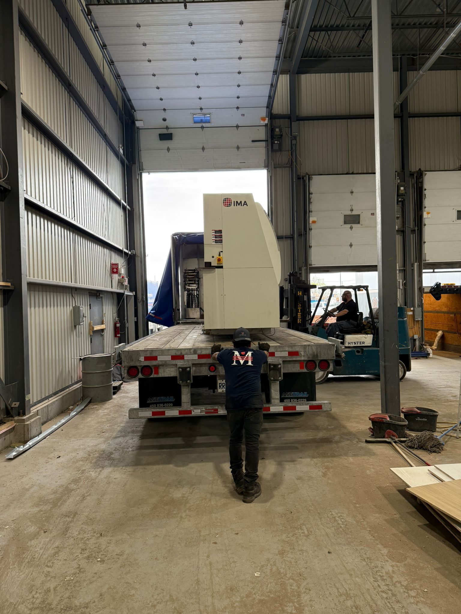 Person guides machine off flatbed truck into warehouse.  Forklift operator in background.