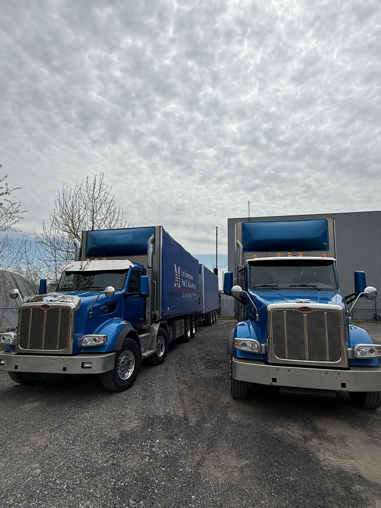 Two blue semi-trucks parked on gravel. Cloudy sky.
