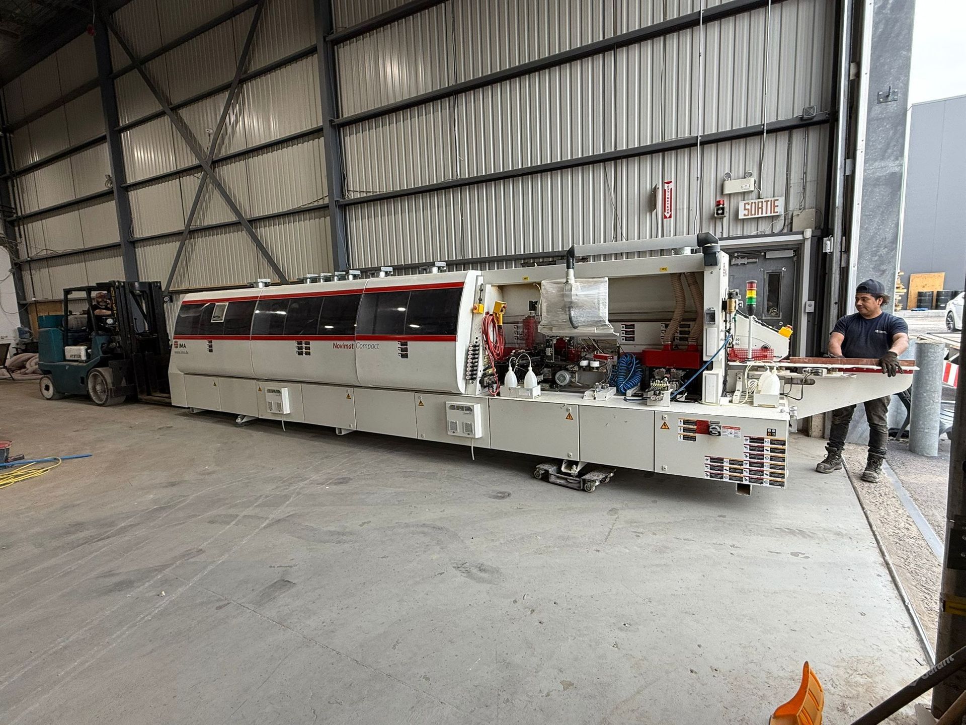 Large white industrial machine being moved by a worker; forklift to the left, inside a warehouse.