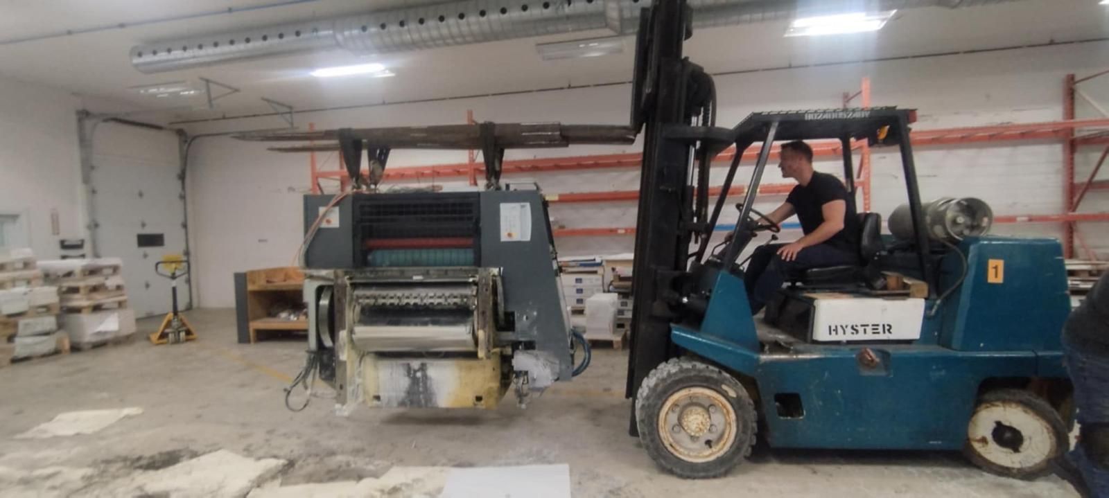 A man driving a blue forklift lifts a large machine in a warehouse.