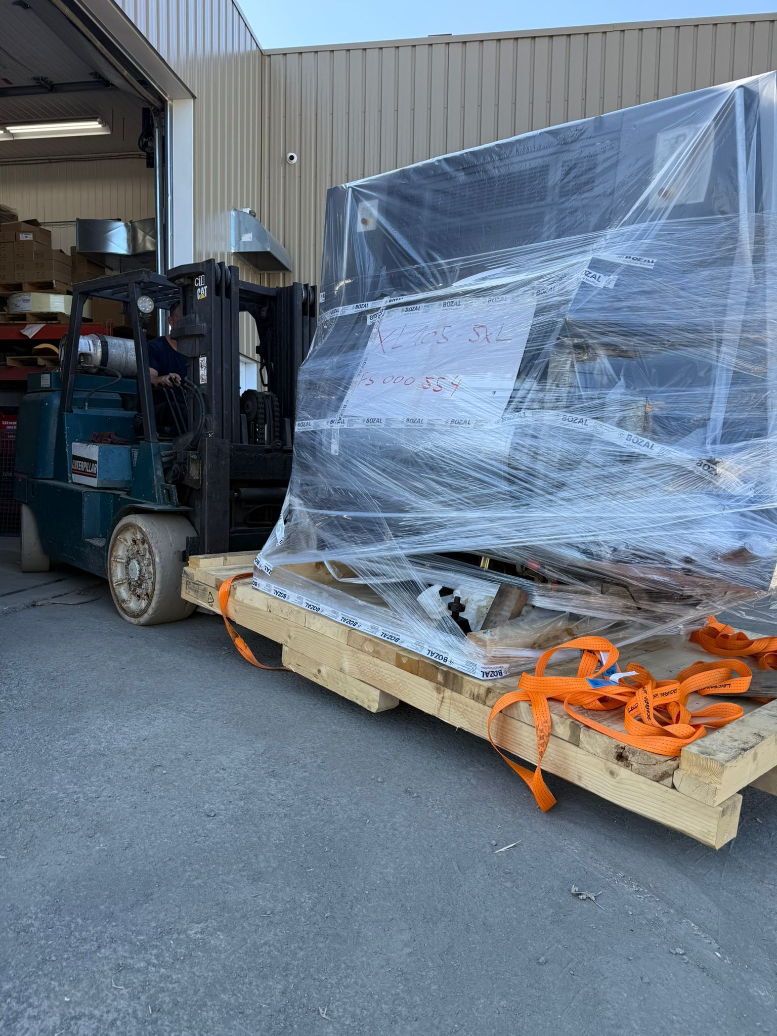 Forklift carrying a large, wrapped object on a wooden pallet, outside a warehouse. Orange straps secure the load.