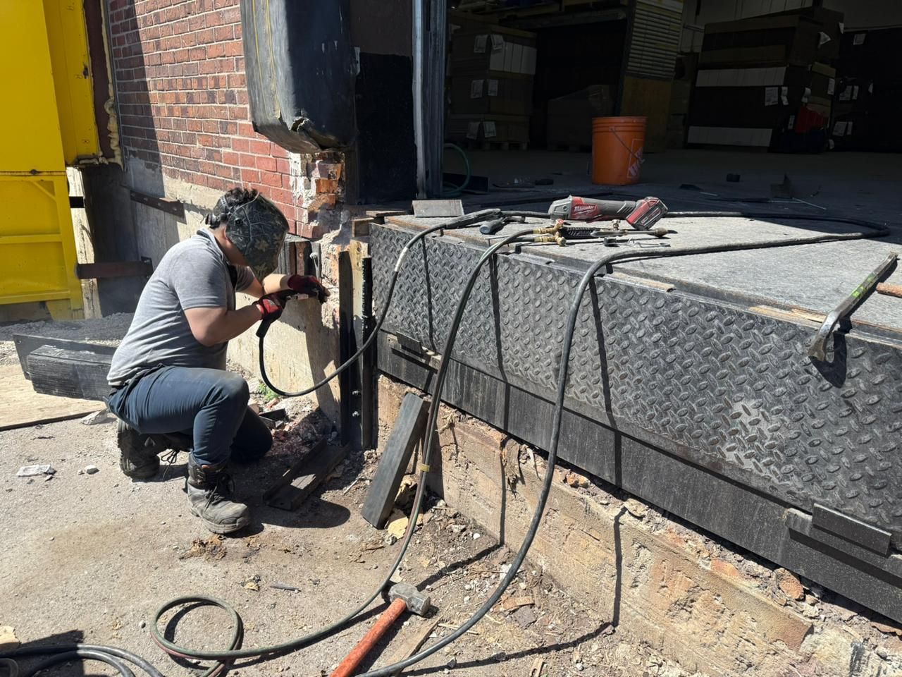 Person welding metal at a loading dock; outdoors, brick wall, protective gear, sparks.