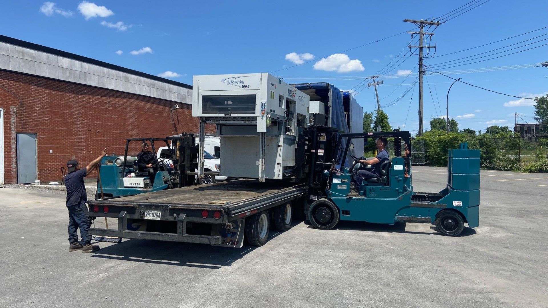 Two forklifts loading large machinery onto a flatbed truck in a parking lot.