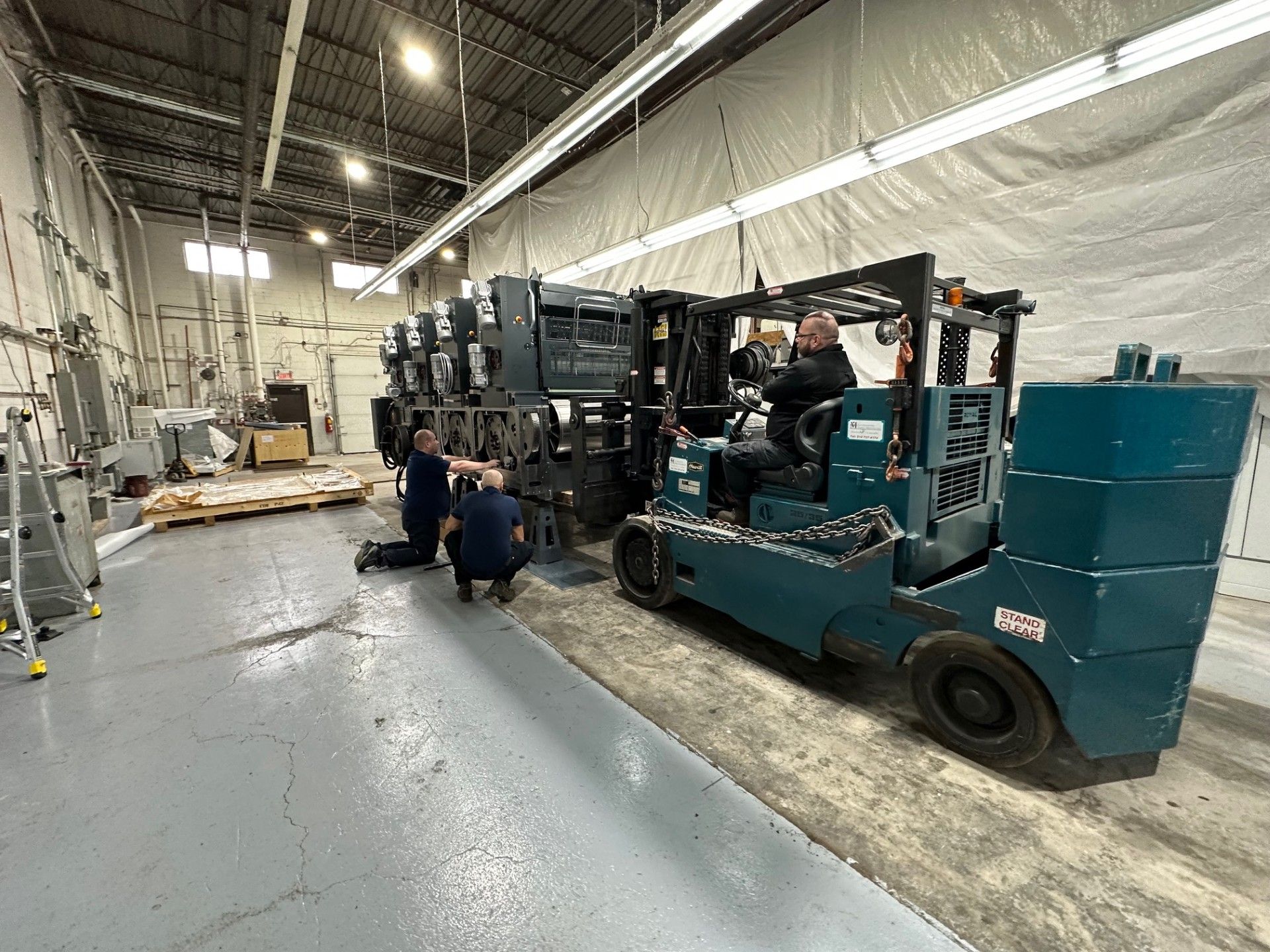 Workers using a forklift to move a large printing machine inside a warehouse.